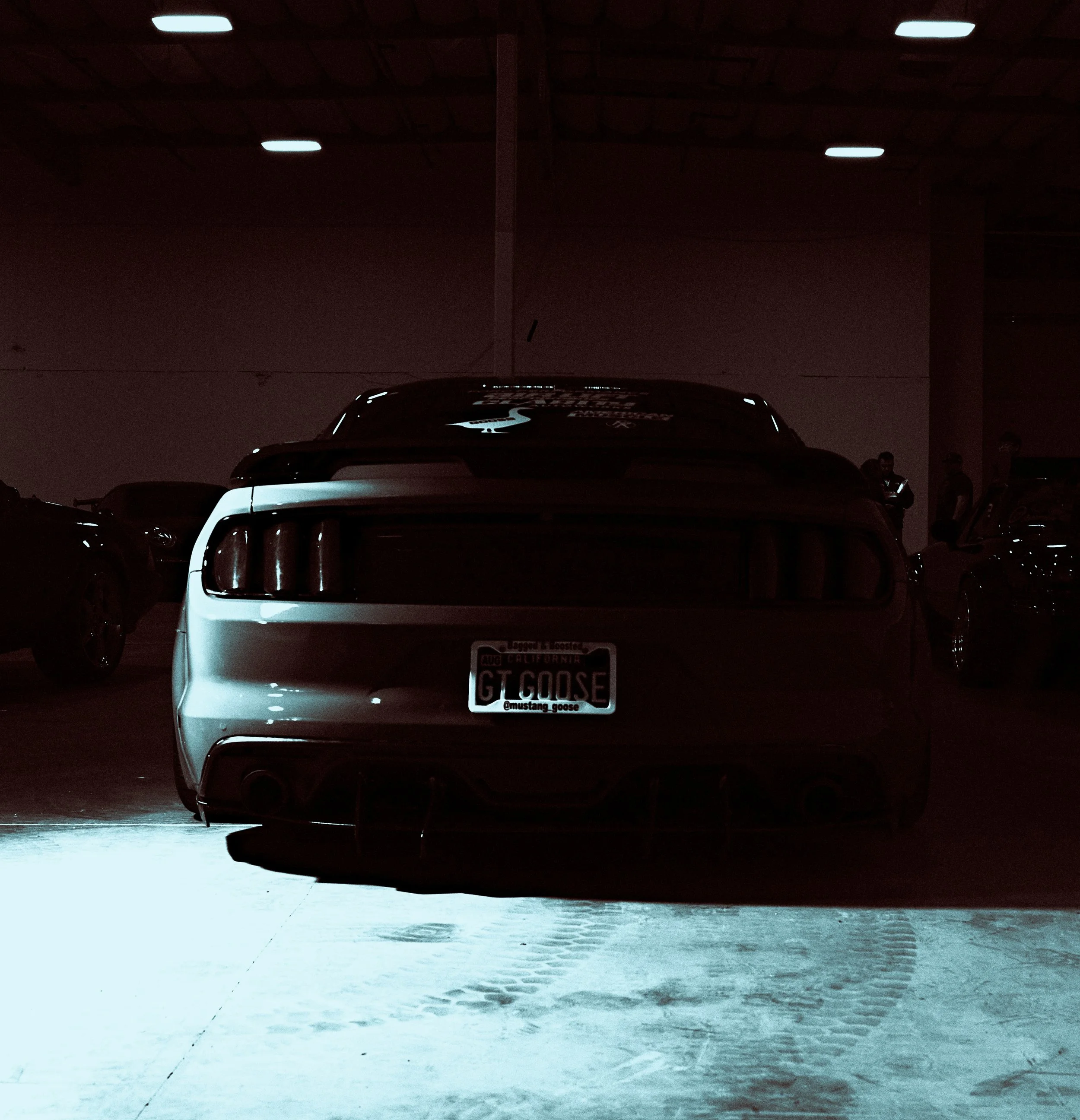 Rear view of a white sports car with a California license plate reading 'GT GOOSE' parked in a dimly lit indoor parking garage, with other cars and a few people in the background.