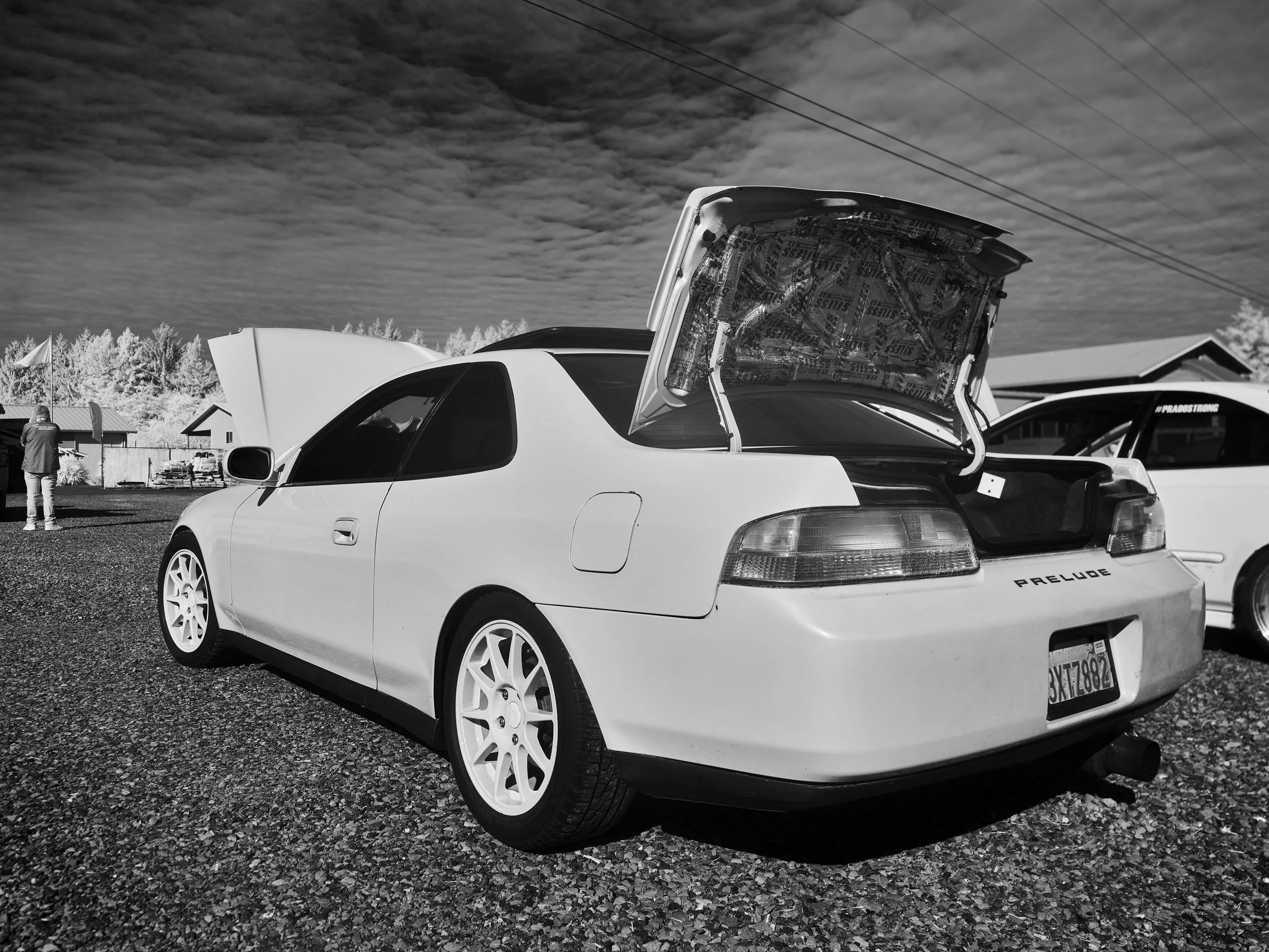 A white sports car with an open rear trunk and front hood, parked outdoors on a gravel surface, with a man and other vehicles and buildings in the background under a cloudy sky.