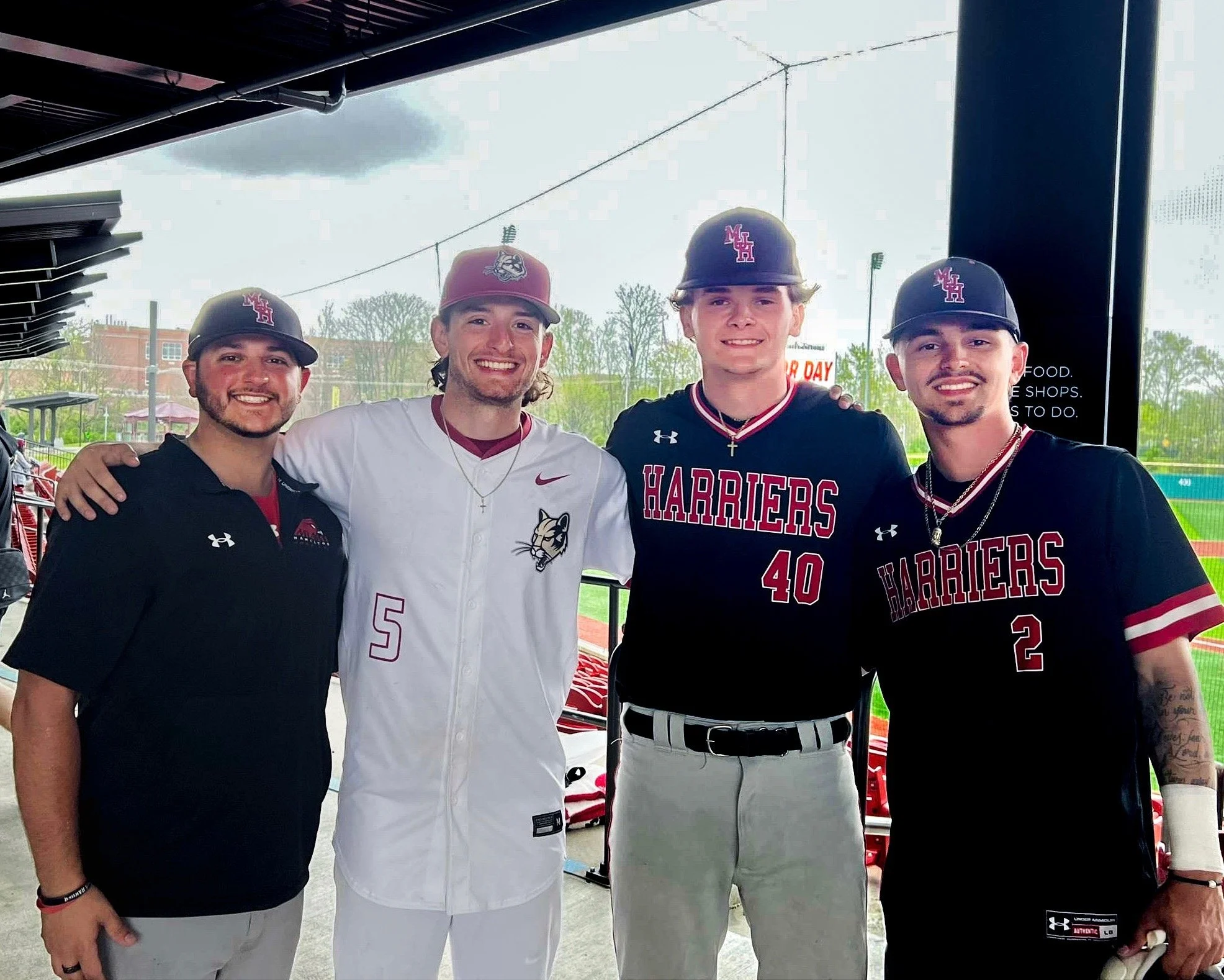 Four young men in baseball uniforms and caps, smiling and posing together at a stadium.