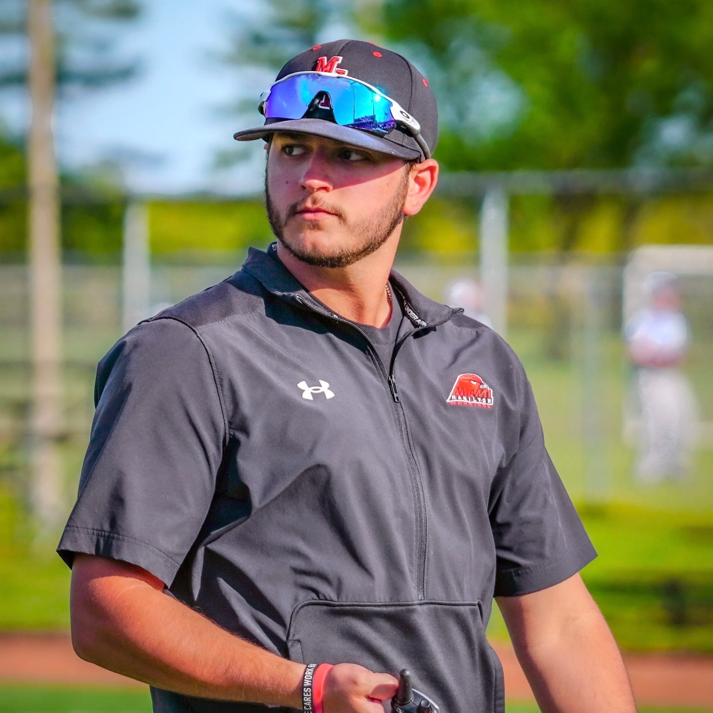 A young man wearing a black baseball cap with red and white logo, reflective sunglasses, and a black jacket with team logos, standing outdoors on a sunny day at a sports field.