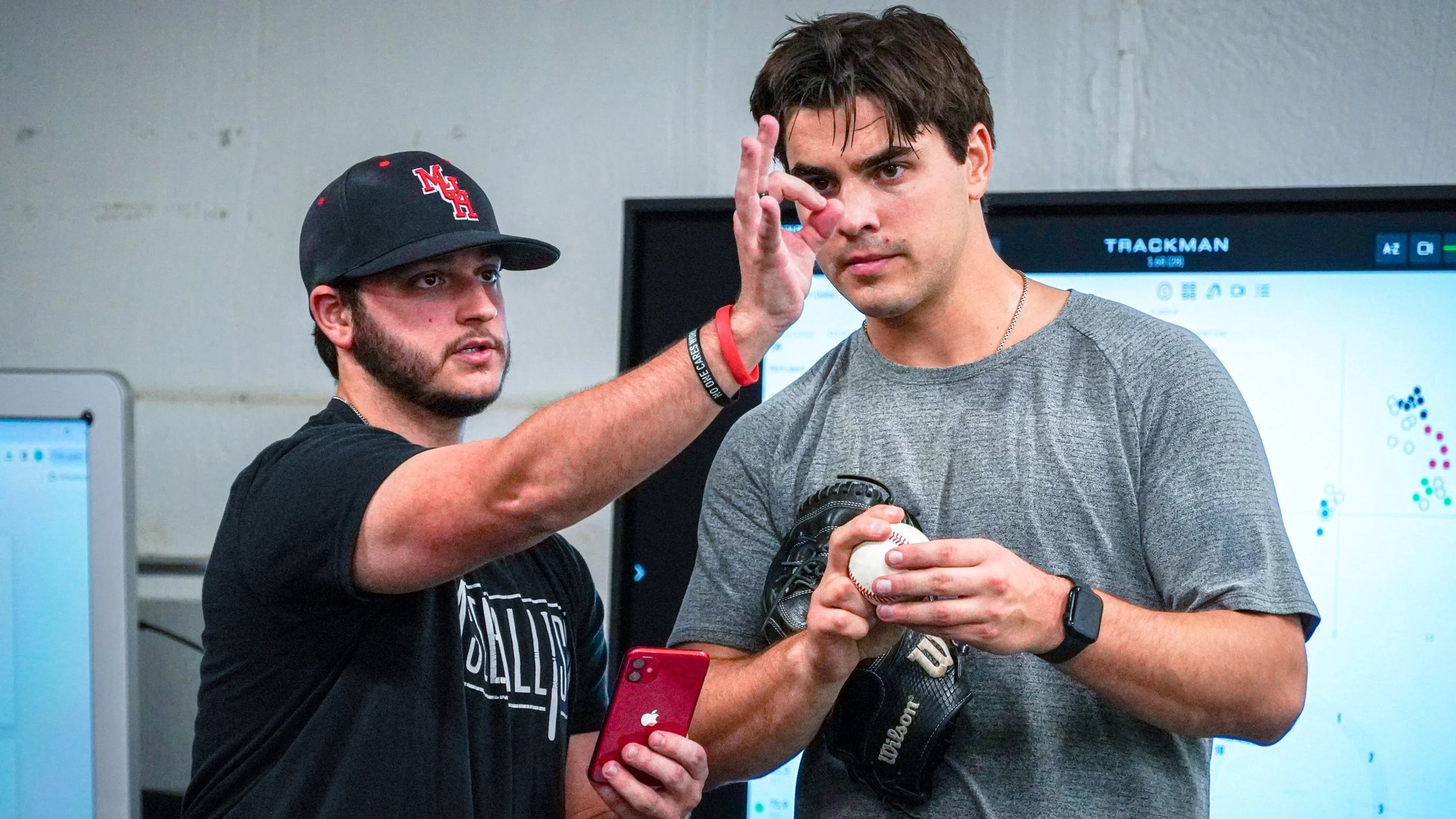 Two men, one in a black cap and shirt and the other in a gray shirt, standing indoors with a wall and screens behind them. The man in the gray shirt is holding a baseball, wearing a glove, and has a wristwatch. The man in the black cap is pointing to the other man's face with his finger.