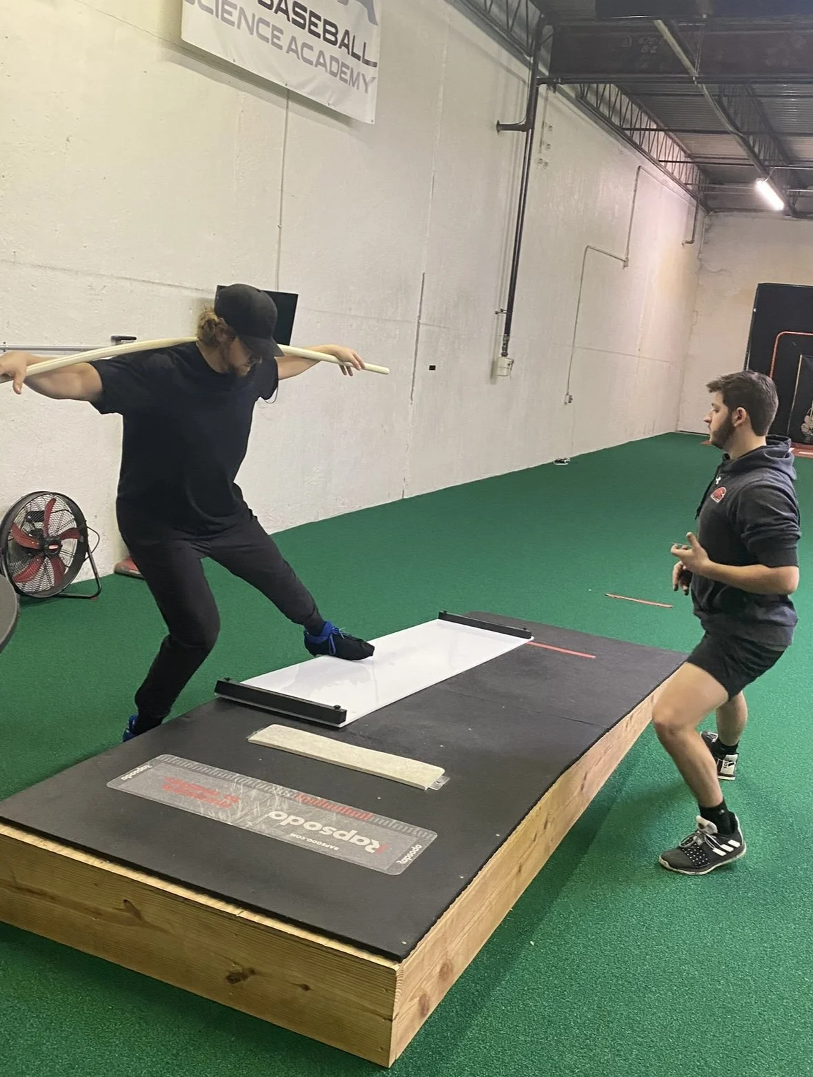 A person practicing baseball in a science academy, mimicking a batting stance with a stick behind their shoulders, while another person watches. The setting is an indoor training facility with green artificial turf and a wooden platform for batting.