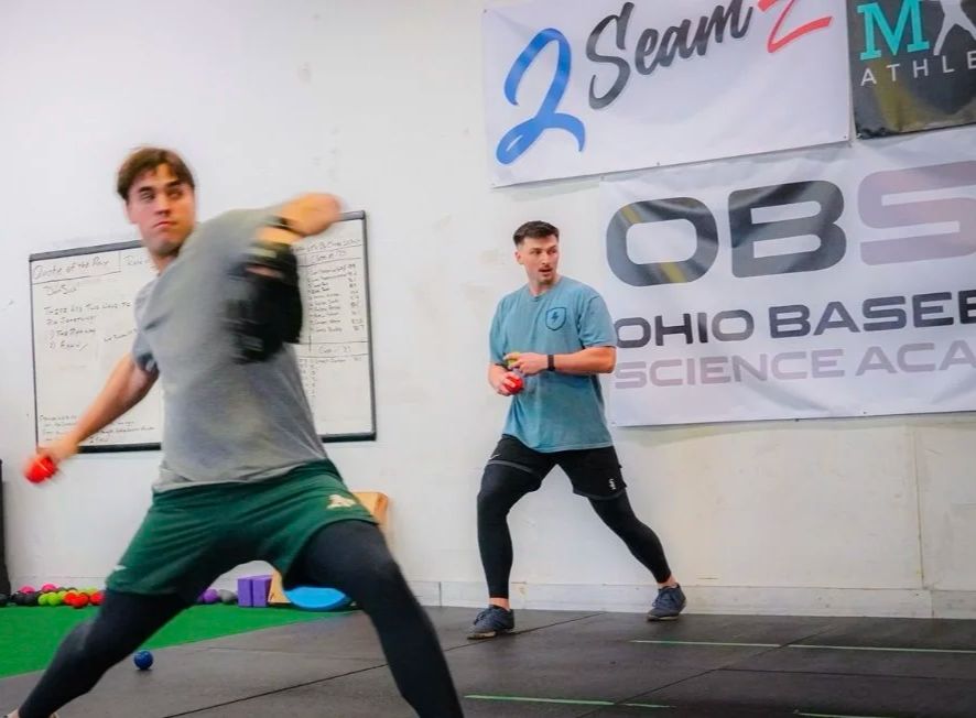 Two men training in a gym with banners on the wall, one in the foreground throwing a ball and another in the background holding a ball