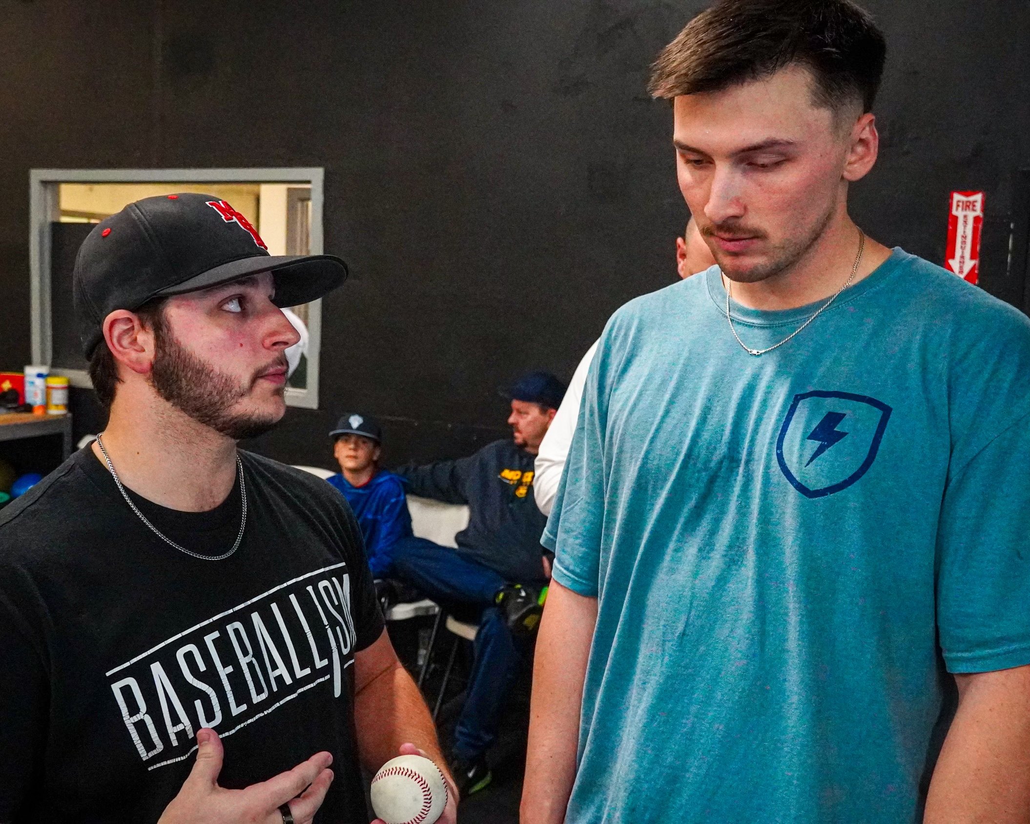 Two young men having a serious conversation in a gym, with other people sitting in the background.