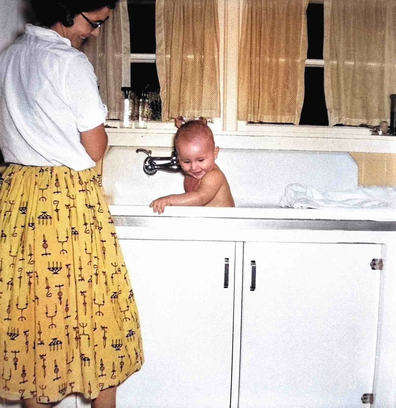 Colorized version of a vintage photo showing a mother and baby at a kitchen sink.