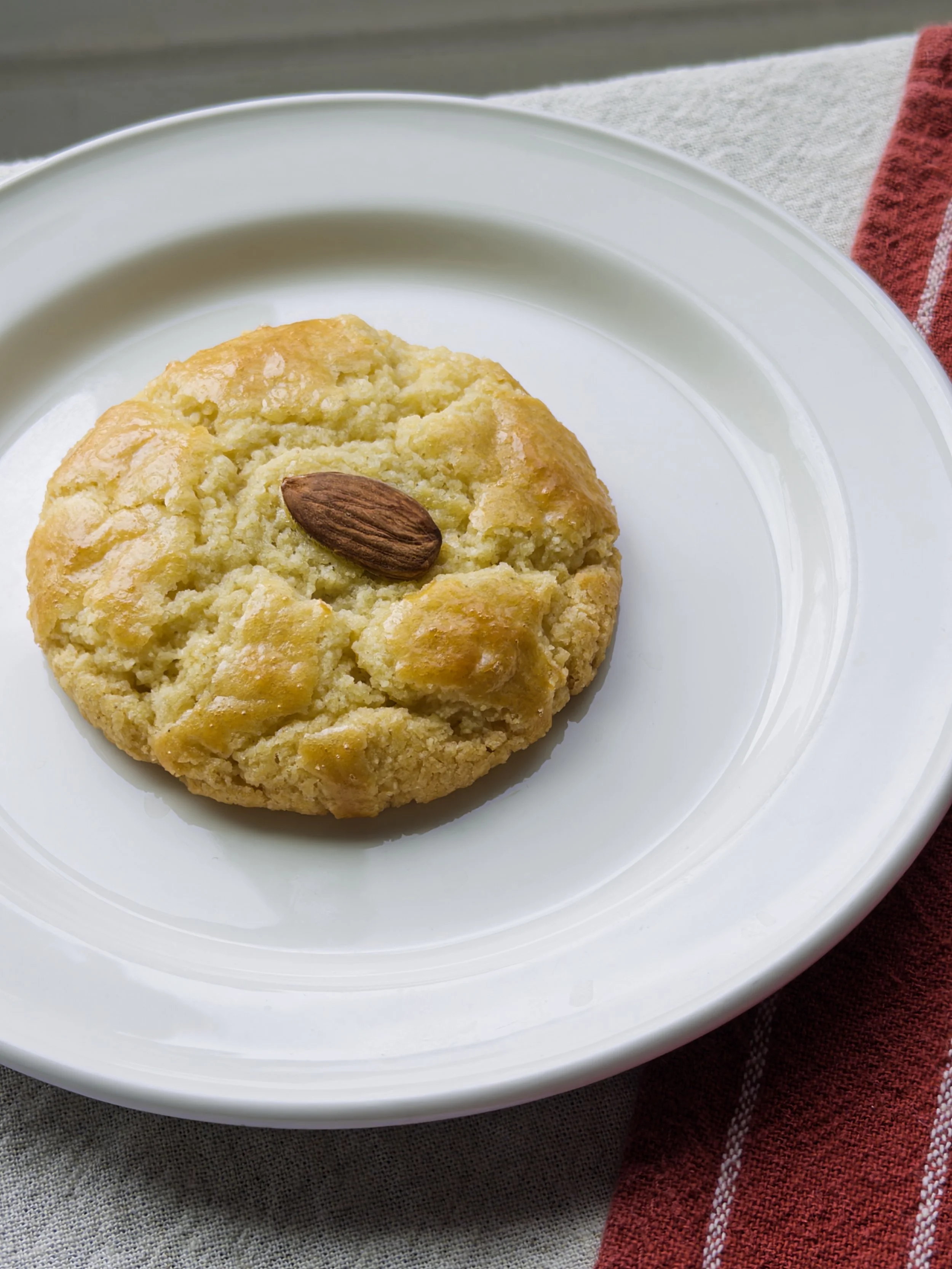 A round, golden-brown cookie topped with an almond on a white plate.