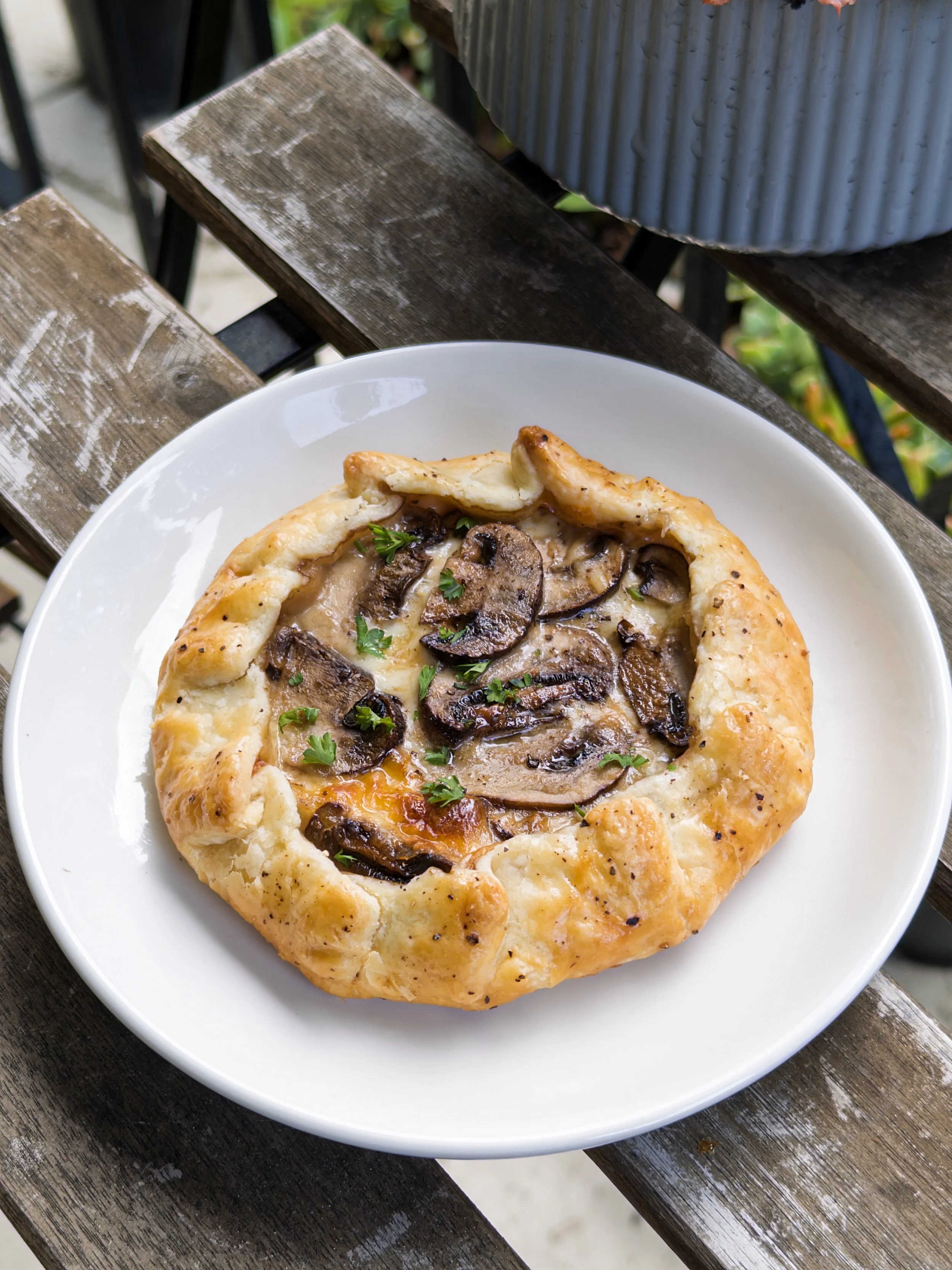 Close-up of a mushroom and cheese galette with herbs on a white plate, placed on a weathered wooden outdoor table.