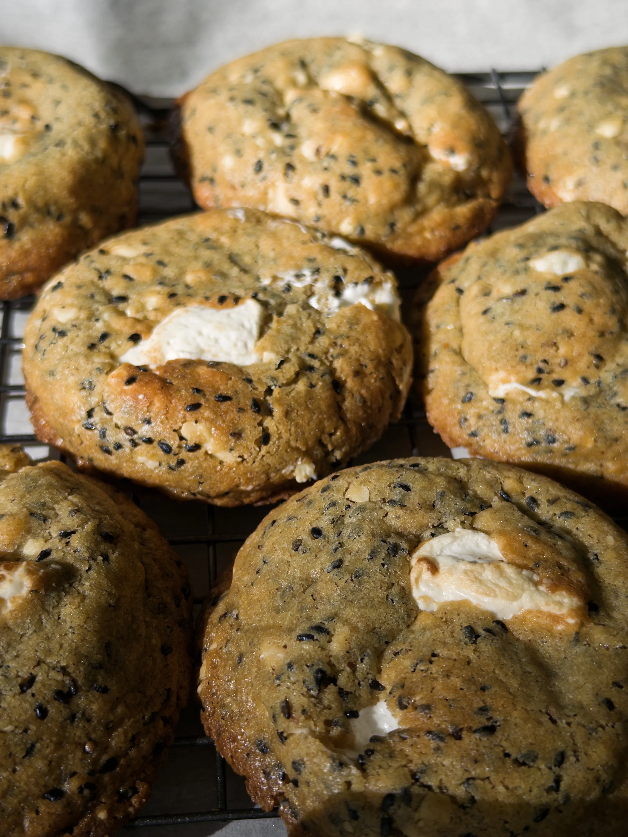 Close-up of freshly baked cookies with white chocolate chunks and black sesame seeds on a cooling rack.