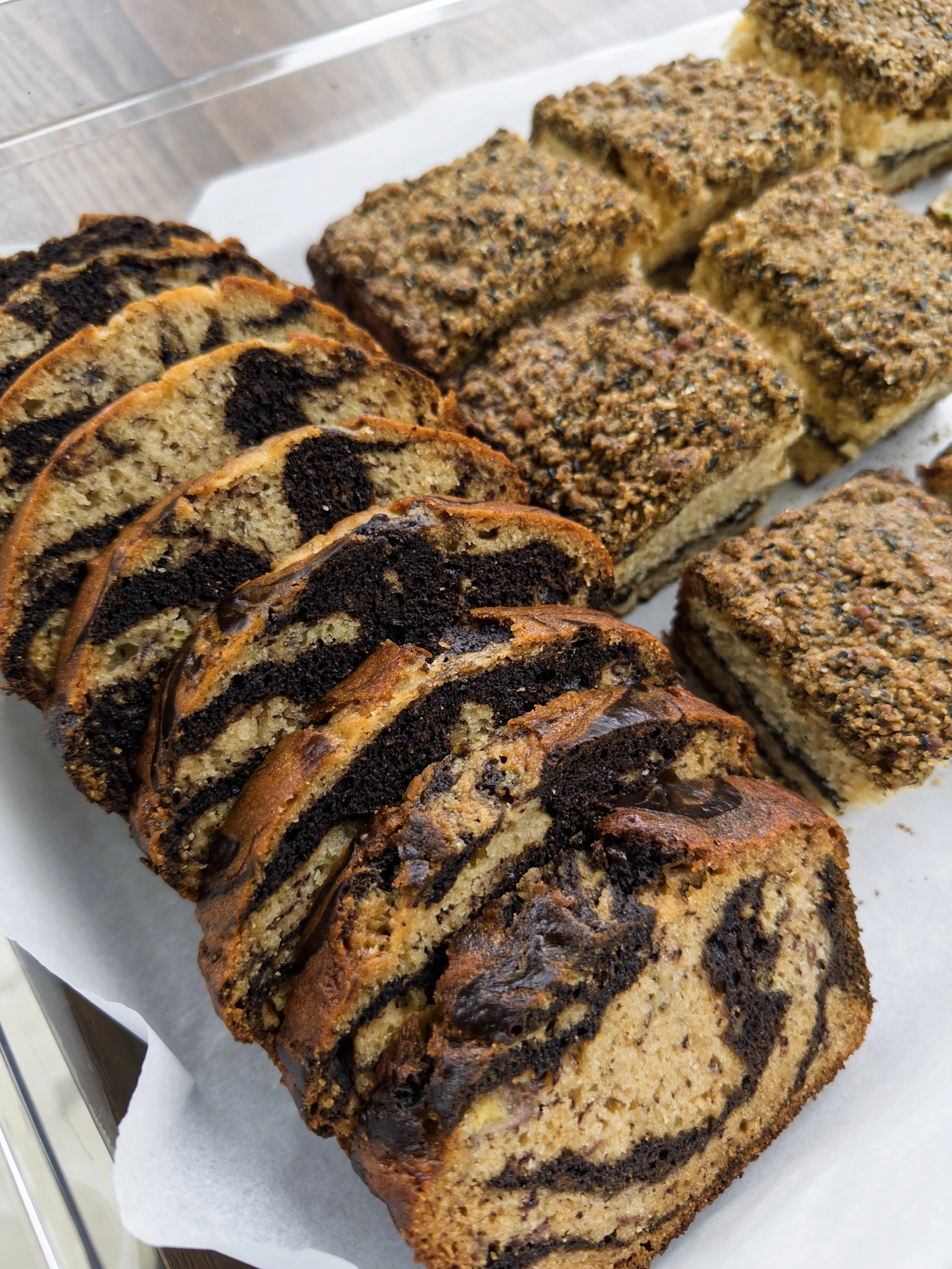 Close-up view of two sliced loaves of banana bread with chocolate drizzle, topped with crushed nuts, placed on parchment paper on a tray.