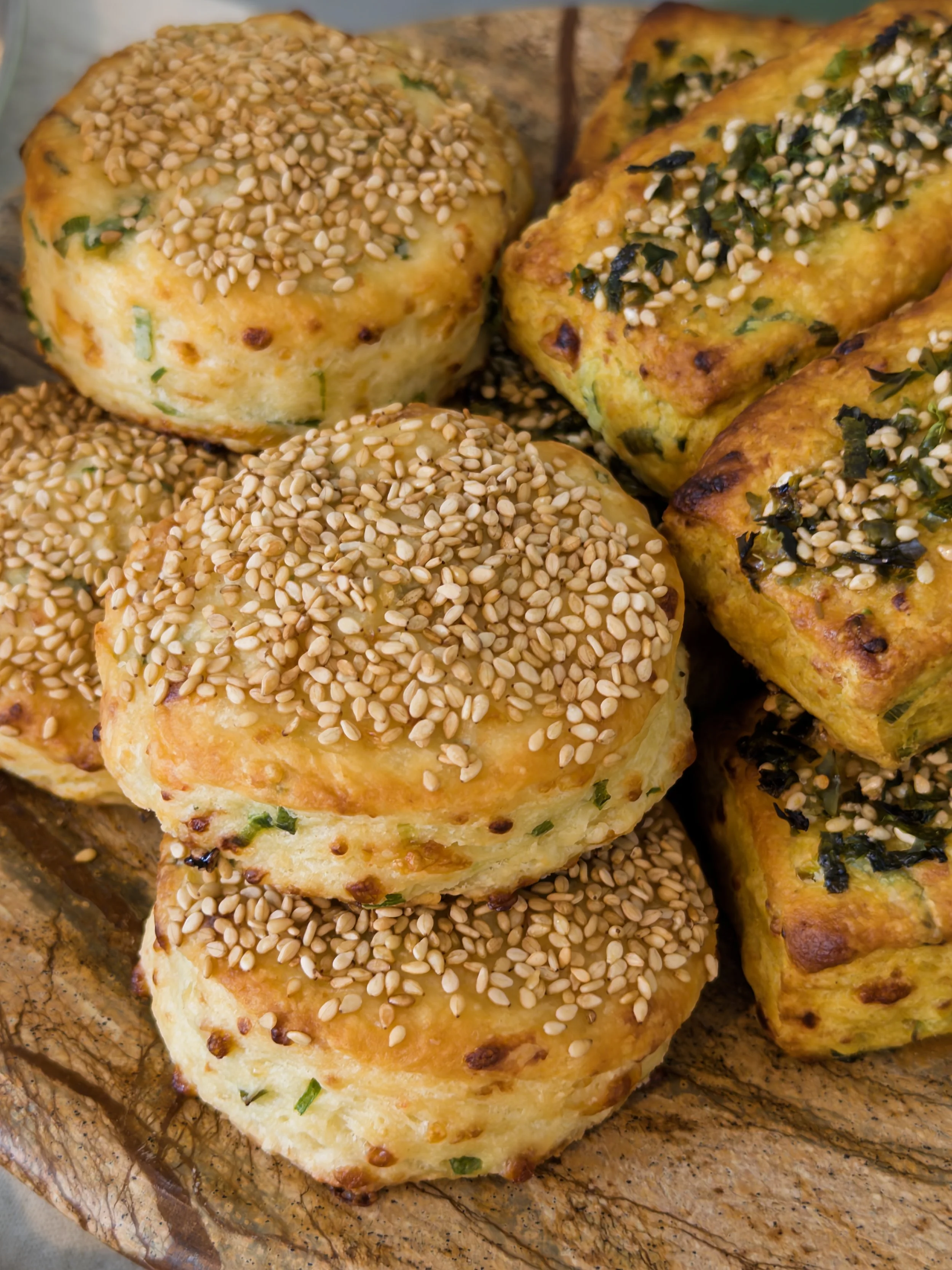 Assorted freshly baked savory bread rolls topped with sesame seeds and chopped herbs on a rustic wooden board.