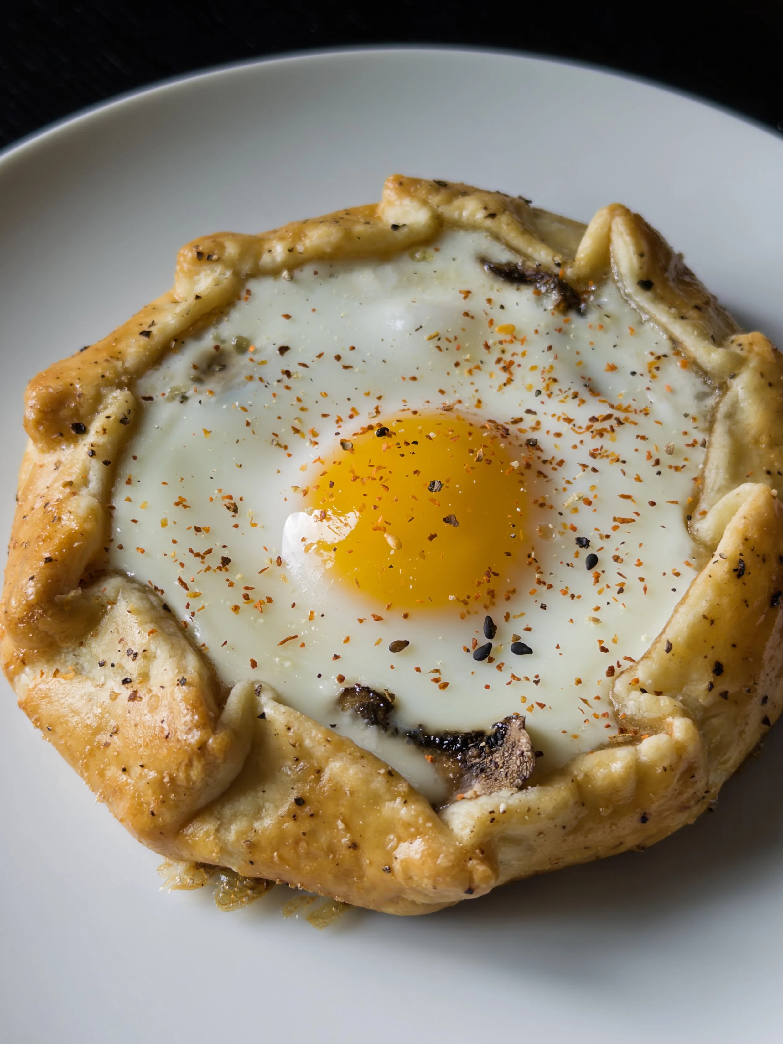 Baked chicken breast with a ring of potato dough crust, topped with a fried egg and sprinkled with black pepper and red pepper flakes, served on a white plate.