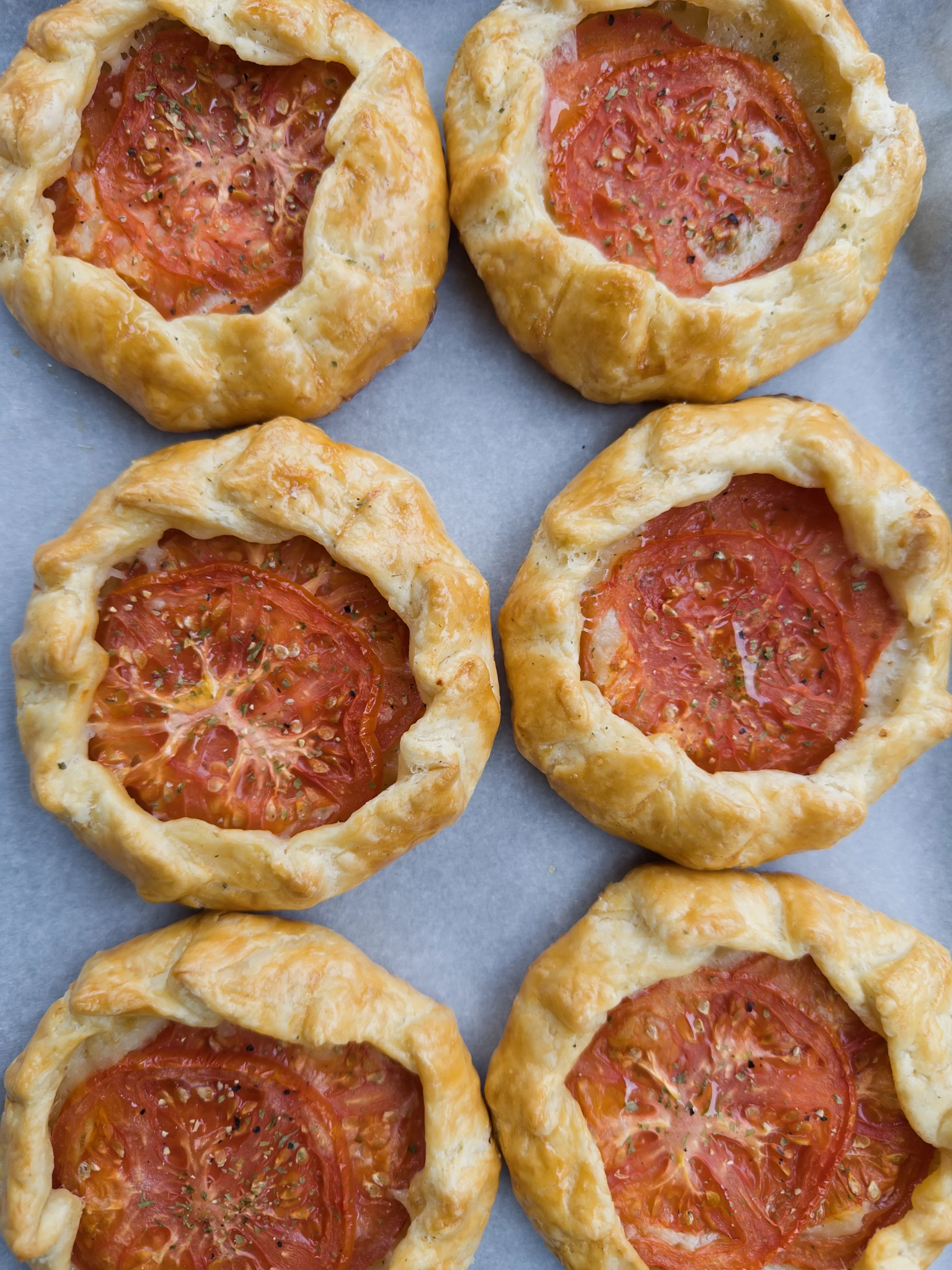 Close-up of freshly baked tomato galettes with golden-brown crust, sliced tomatoes, herbs, and sprinkled spices on top.