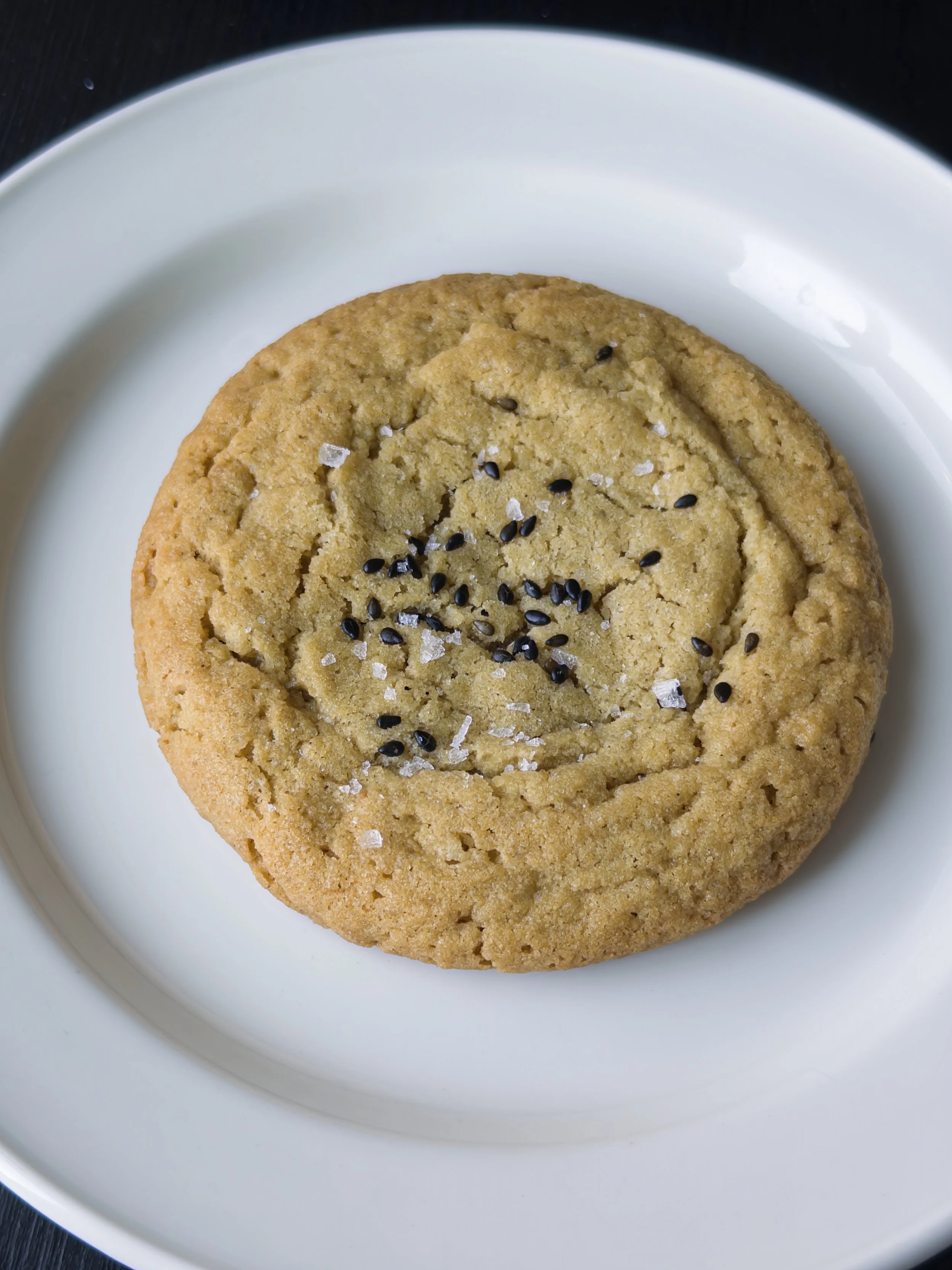 Close-up of a large chocolate chip cookie with black sesame seeds and coarse salt on top, placed on a white plate.