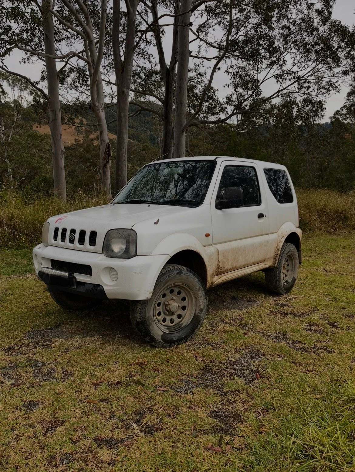 A white Suzuki SUV with dirt on the lower sides, parked on grass near trees.