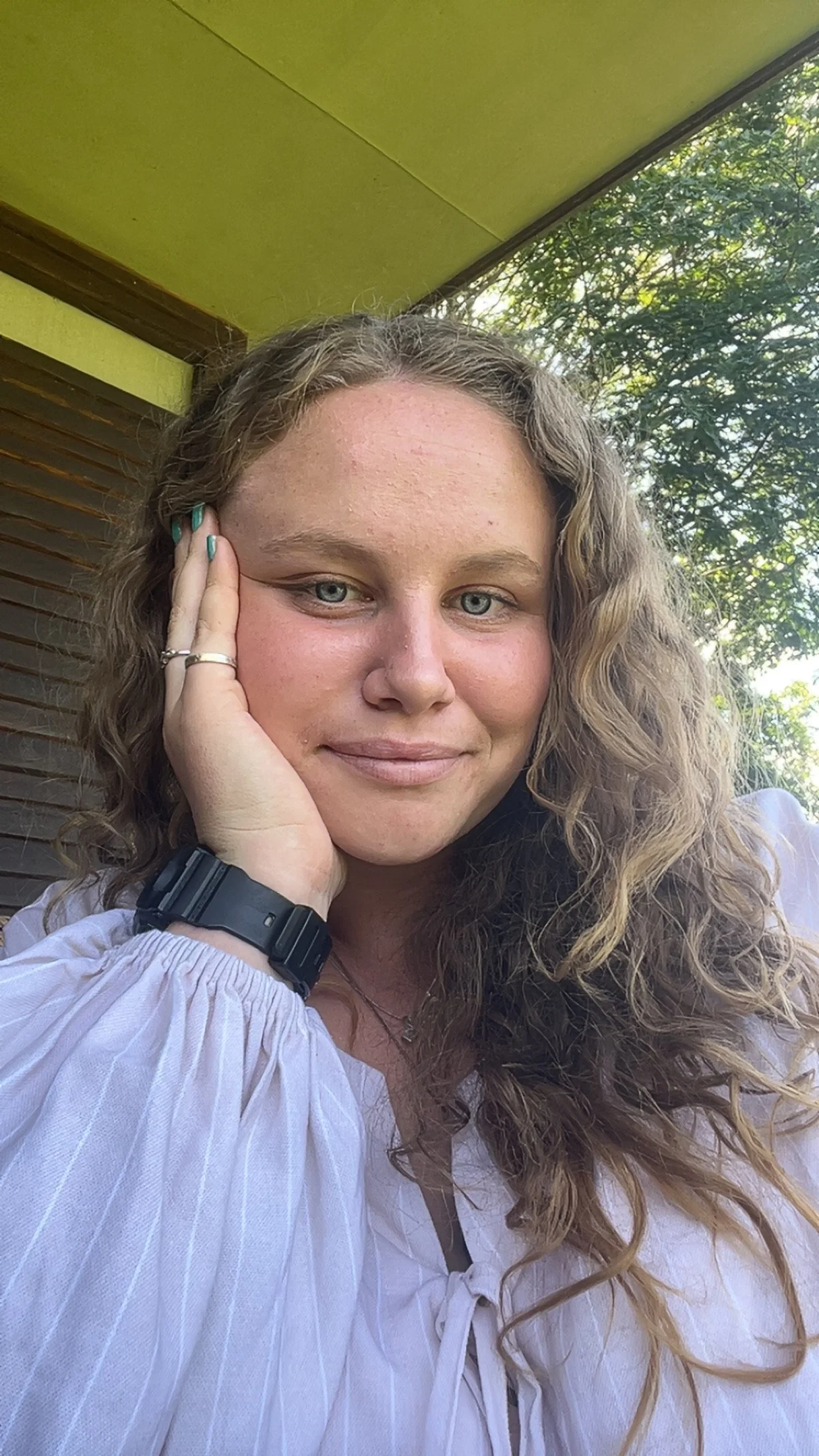 A woman with light skin and long, curly blonde hair, resting her face on her hand, smiling softly outside near greenery.