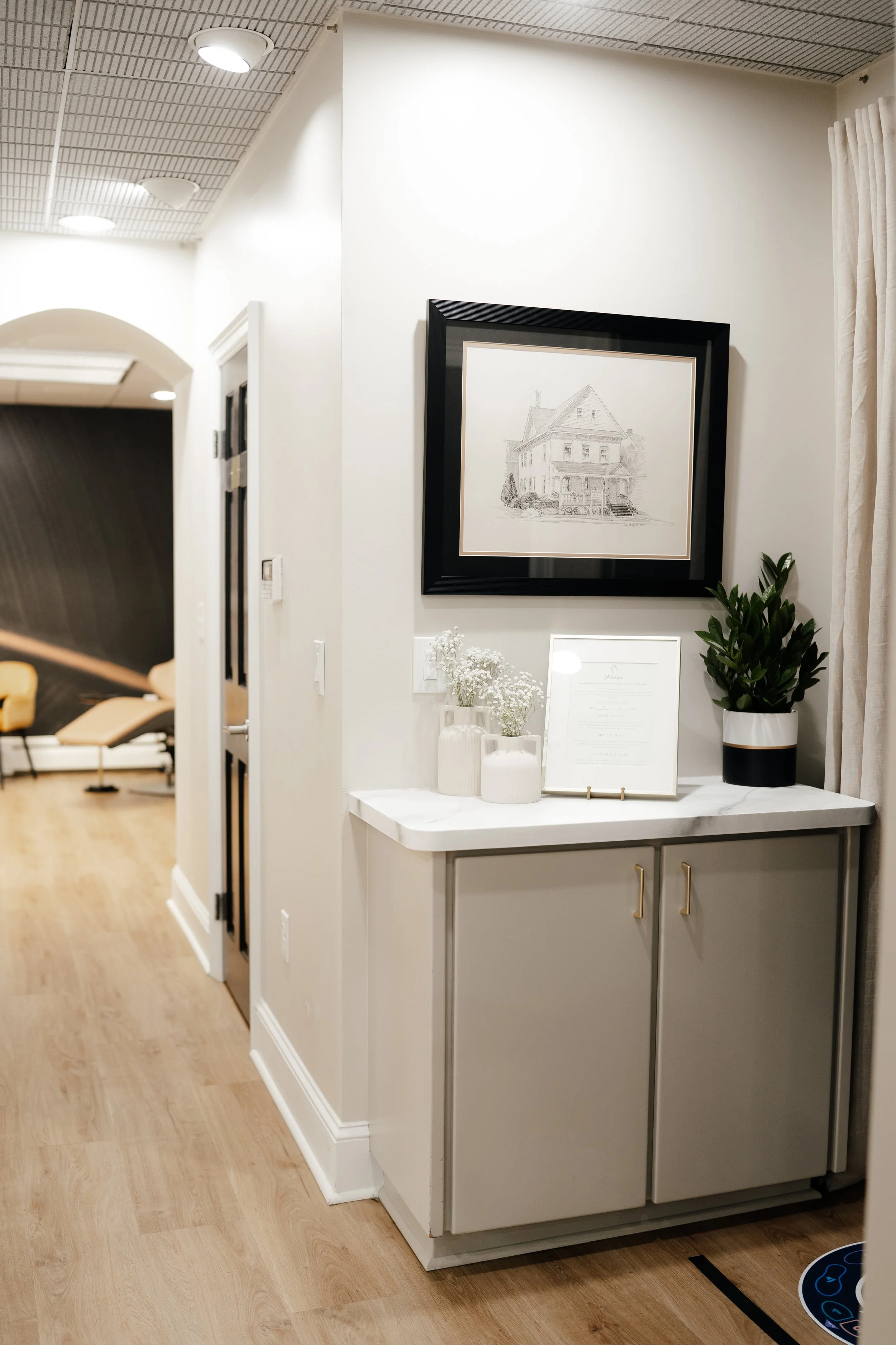 A hallway corner with a white cabinet topped with flower vases, a framed document, and a potted plant; a framed drawing of a house hangs above the cabinet; soft lighting and curtains on the right.