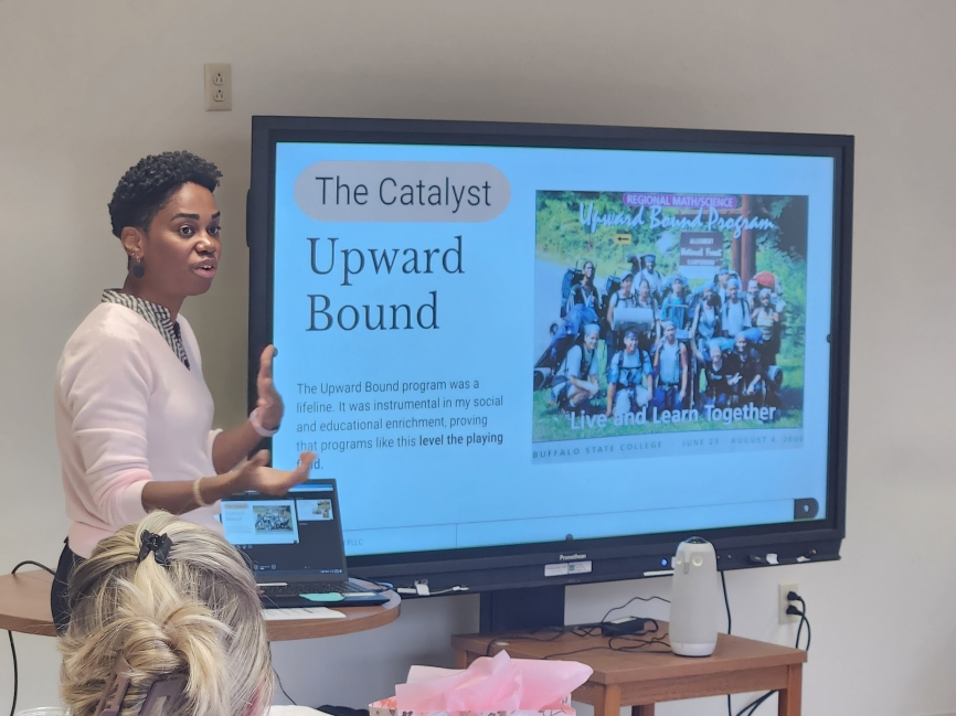 A woman giving a presentation in front of a large screen displaying information about the Upward Bound program at Buffalo State College. The woman is gesturing with her hands while several people are seated and listening.