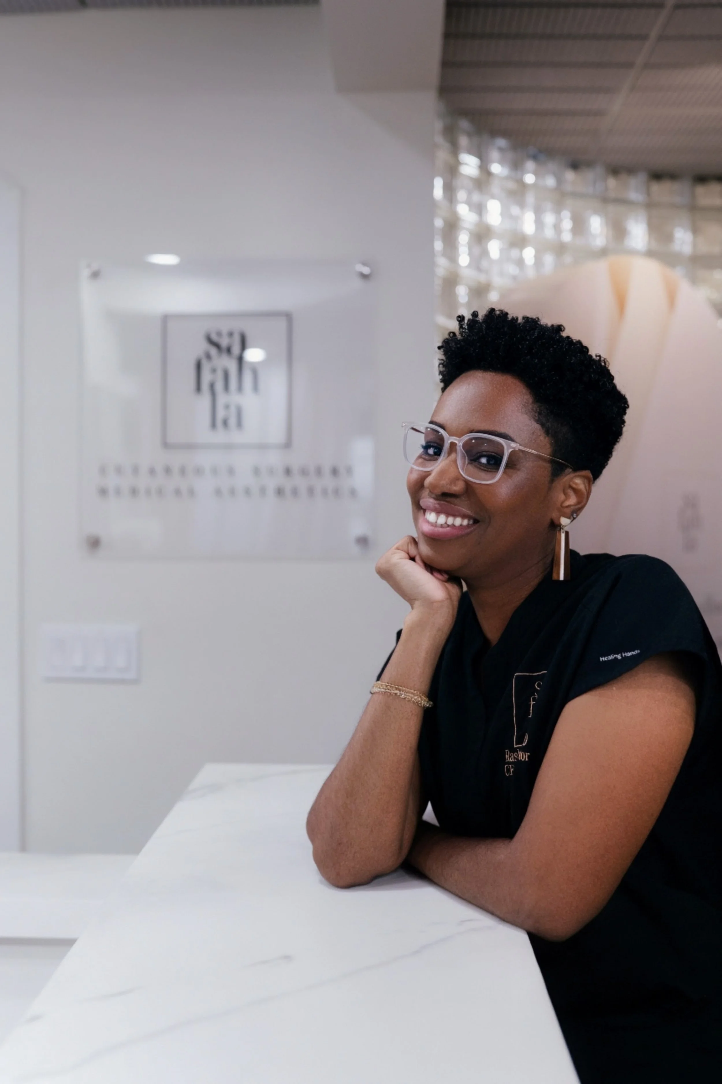 Dr. Rashida Vassell with short curly hair, glasses, and earrings smiling and resting her chin on her hand at a white marble counter, in front of a blurred wall with a logo sign.