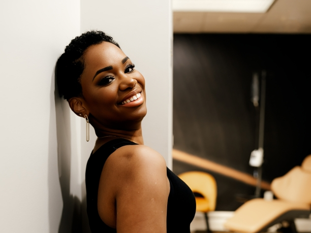 A young woman with short curly hair smiling and leaning against a wall in an indoor setting.
