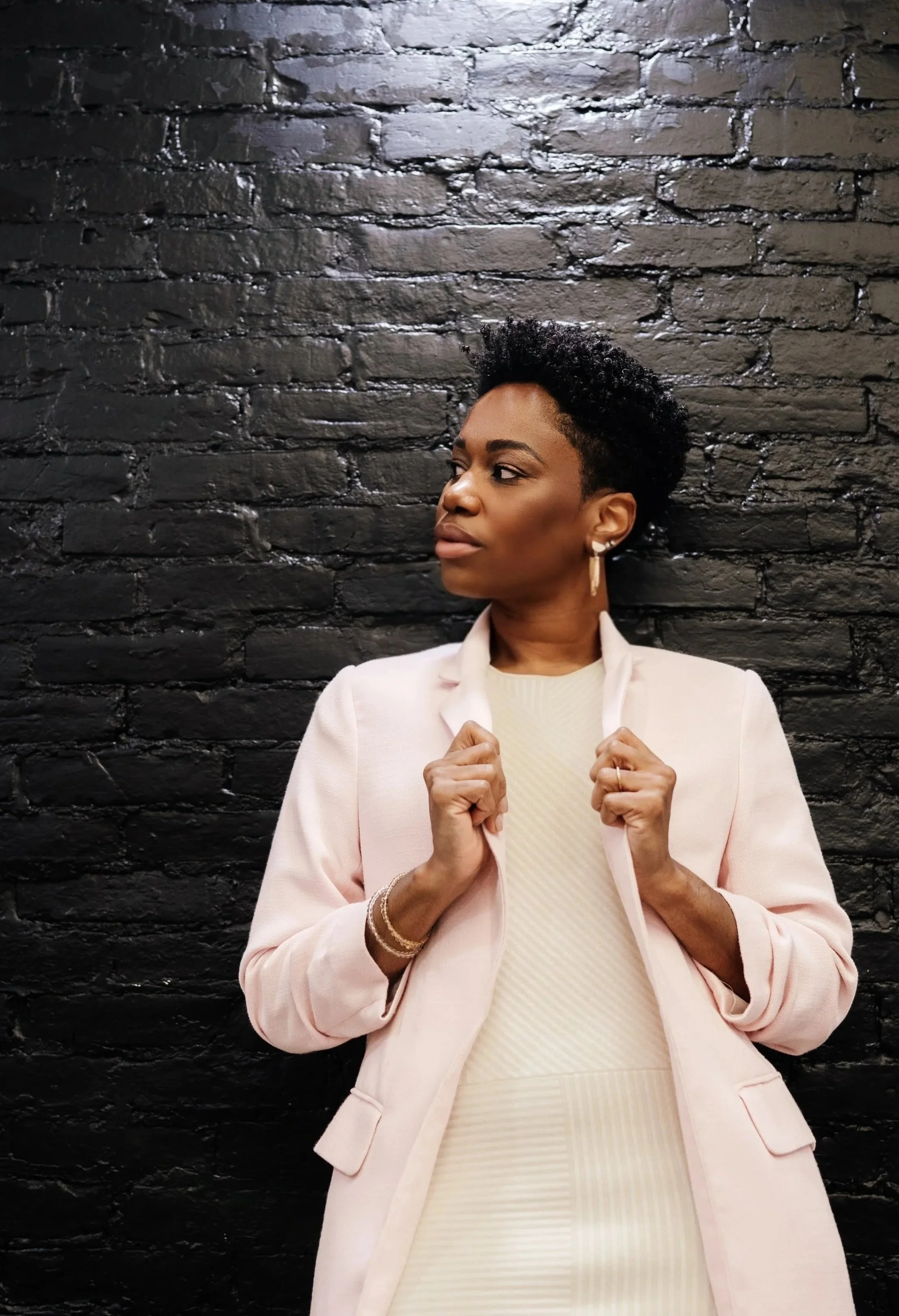 Dr. Rashida Vassell with short curly hair and hoop earrings standing in front of a black brick wall, wearing a light pink blazer over a cream-colored dress, posing confidently.