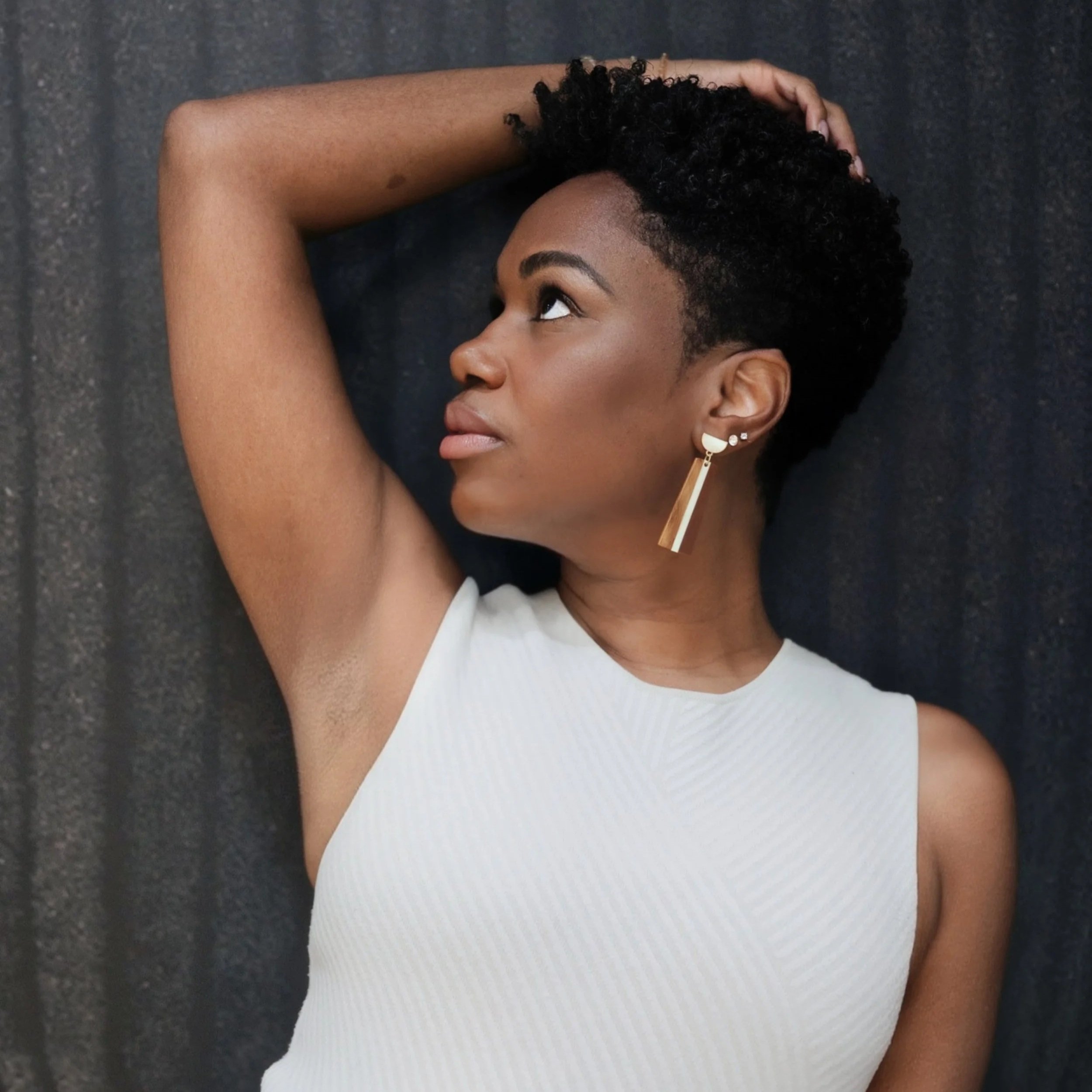 Dr. Rashida Vassell, a young woman with short curly hair, wearing a sleeveless white top and long earring, looks to her left with her right arm raised and resting on her head, standing against a dark background.