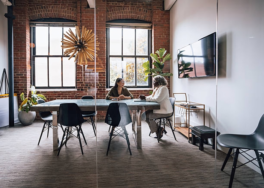 Two women sitting at a conference table in a modern office with exposed brick walls, large windows, potted plants, and a flat-screen TV on the white wall.