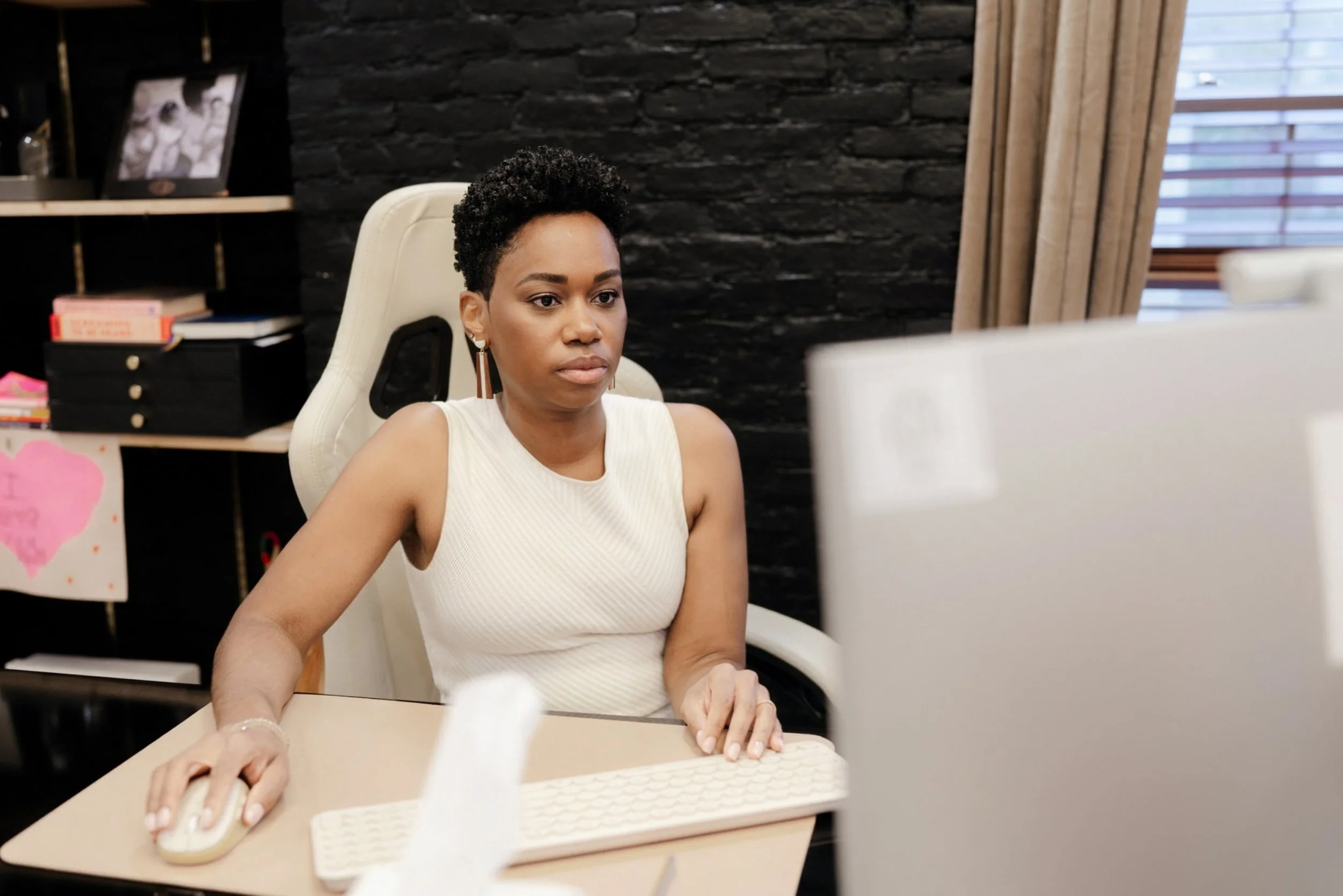 Dr. Rashida Vassell working at a desk, using a computer mouse, in an office with dark brick wall and window with blinds.