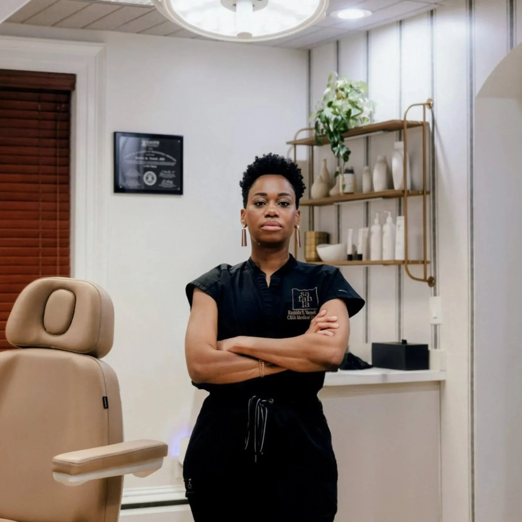 Dr. Rashida Vassell, a confident woman with short curly hair and earrings standing with arms crossed in a spa or skincare clinic, with shelves of skincare products behind her.