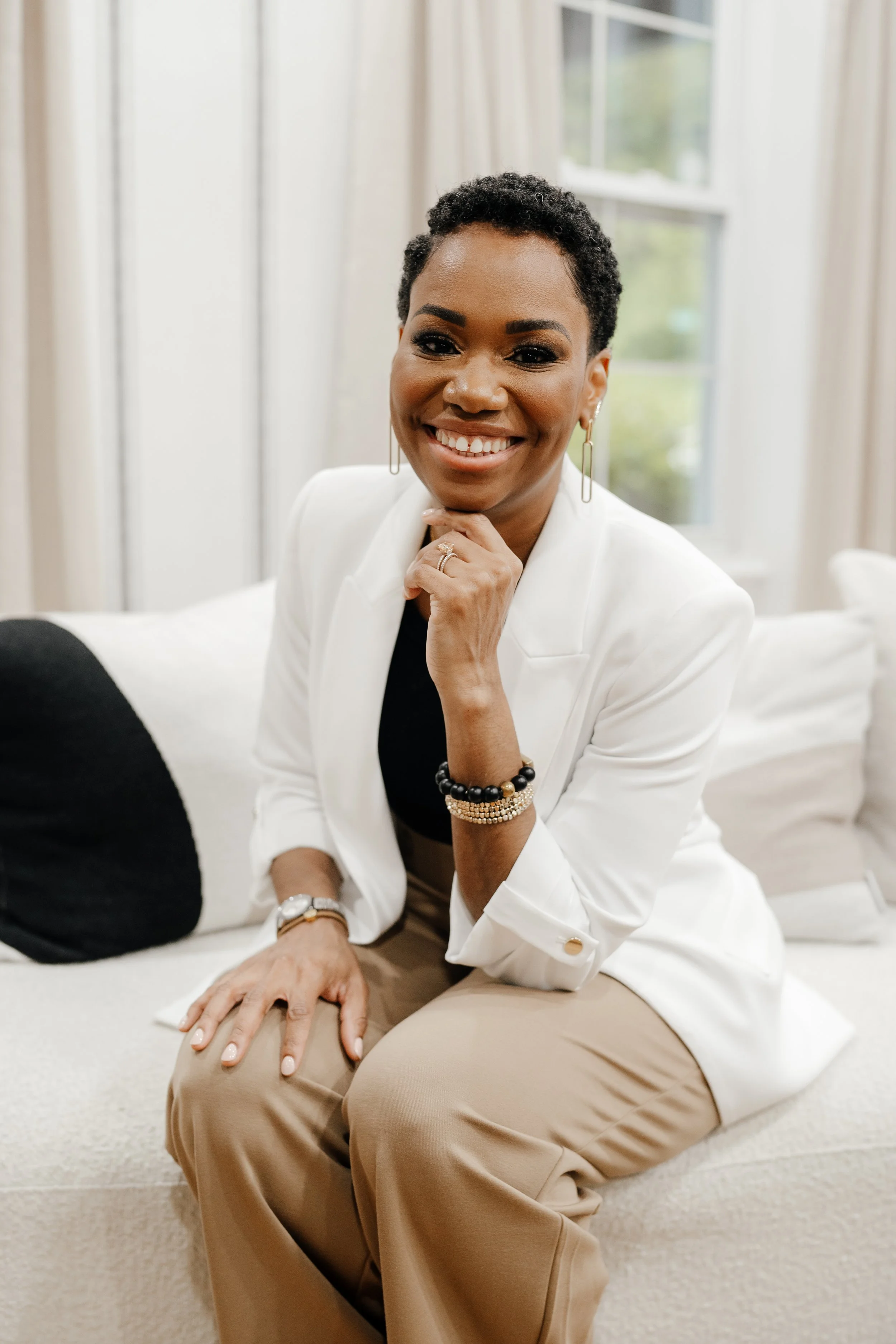 A woman with short curly hair sitting on a white couch, smiling, wearing a white blazer over a black top, beige pants, and various jewelry including earrings, bracelets, and a watch, in a bright room with large windows in the background.