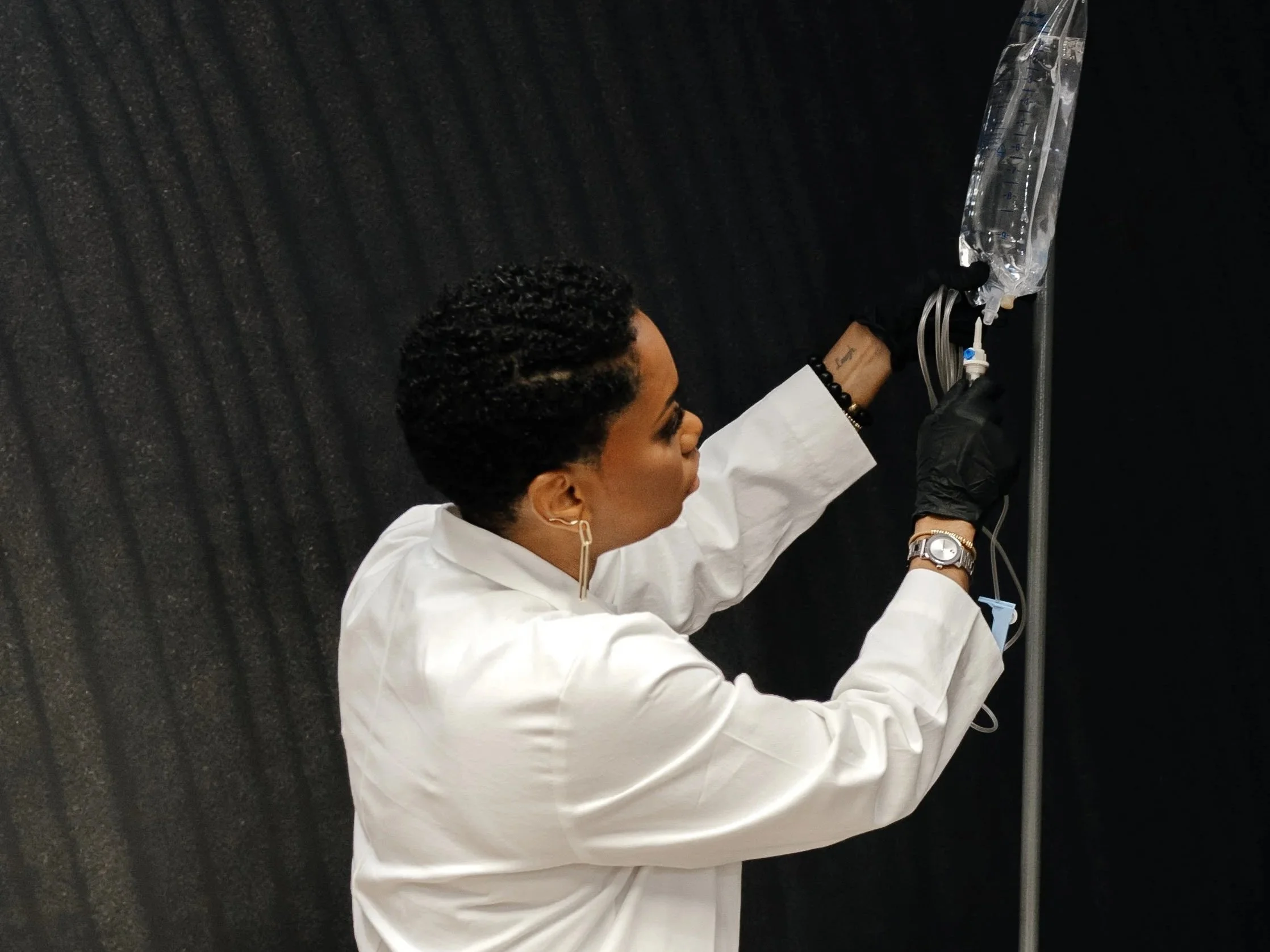 A woman in a white lab coat preparing an IV drip against a black wall.