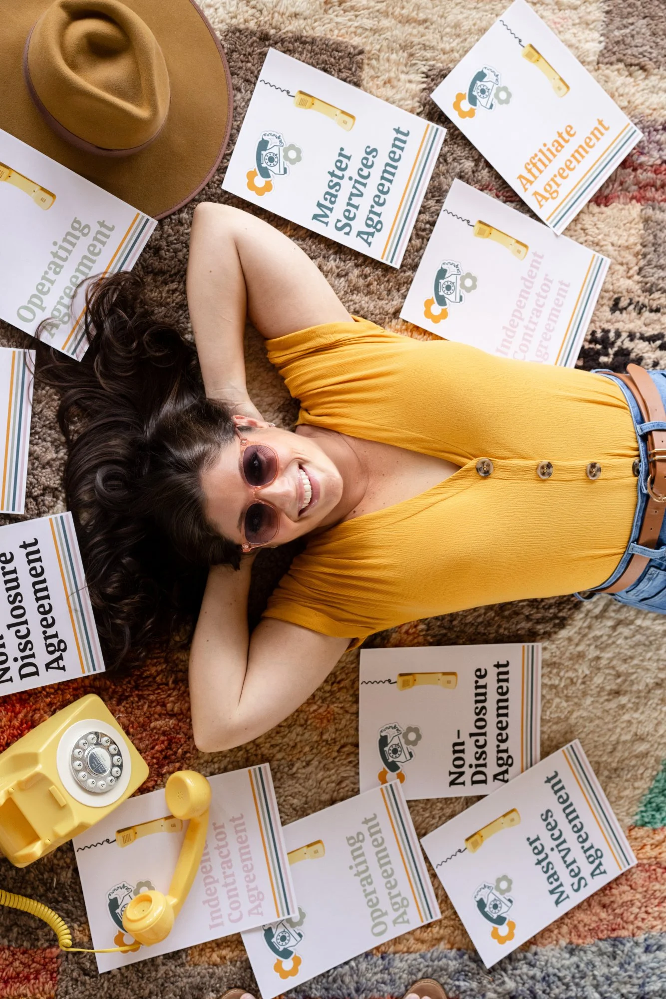 A woman lying on a colorful carpet with her arms behind her head, wearing a yellow top and sunglasses, smiling. She is surrounded by signs that read "Non-Disclosure Agreement," "Master Services Agreement," and "Affiliate Agreement," along with a vintage yellow rotary telephone.