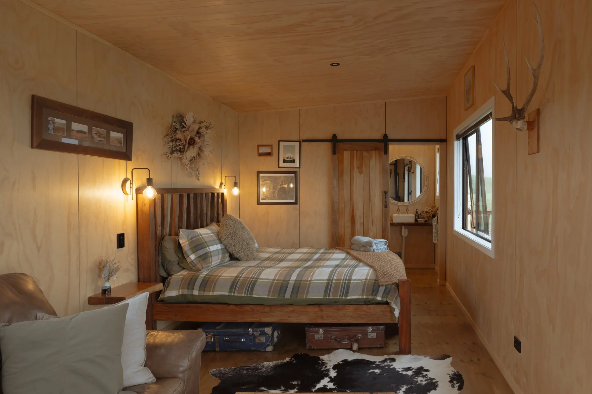 Rustic bedroom at The Farmers Rest with wooden walls and ceiling, featuring a bed with plaid bedding, a leather armchair, and various wall decorations including framed photos, a mirror, and mounted antlers. 