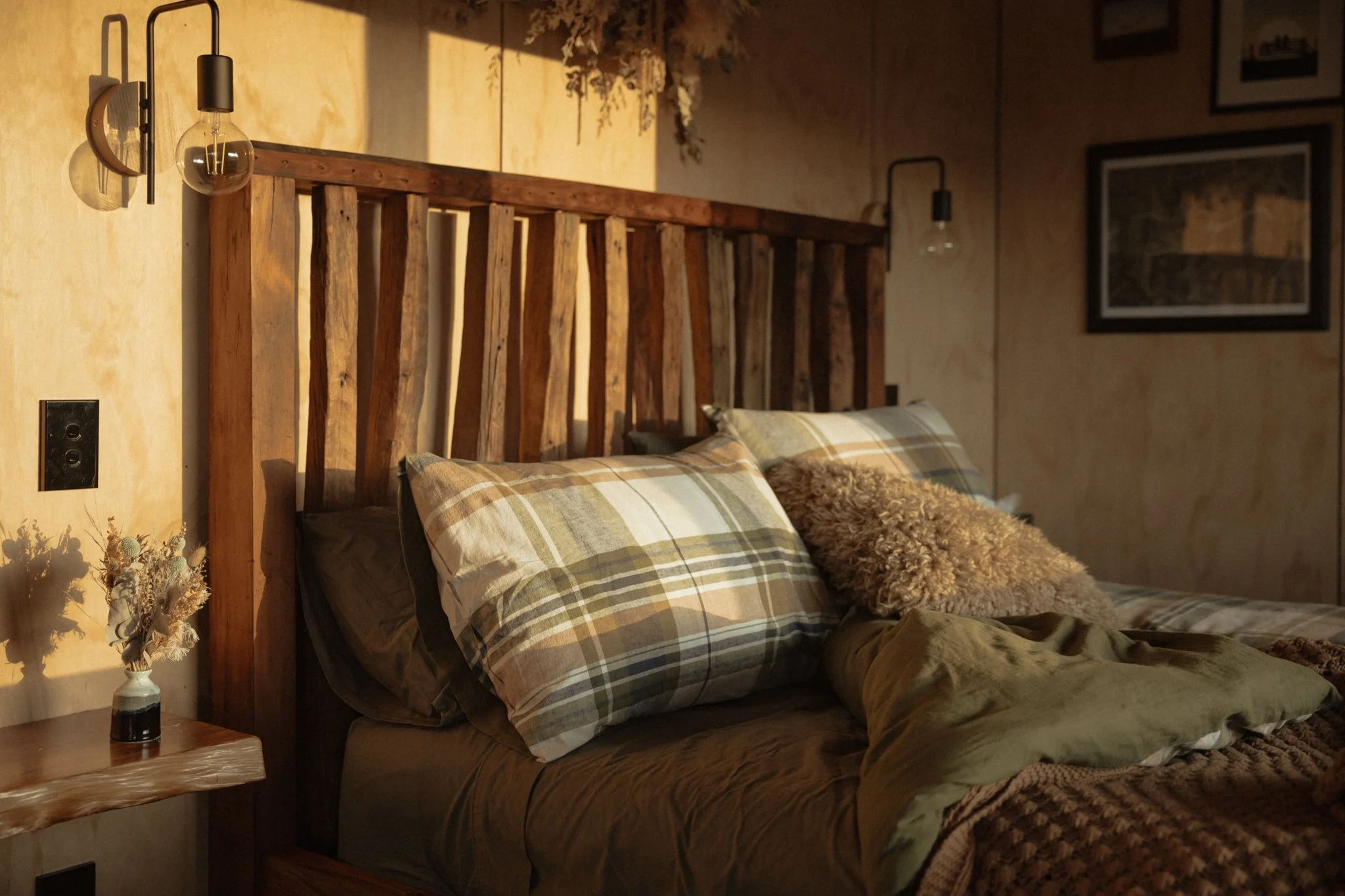 A cozy bedroom with a wooden headboard, plaid pillows, and a small side table holding a dried flower arrangement. Sunlight casts warm shadows on the wall, creating a relaxing atmosphere at The Farmers Rest cabin