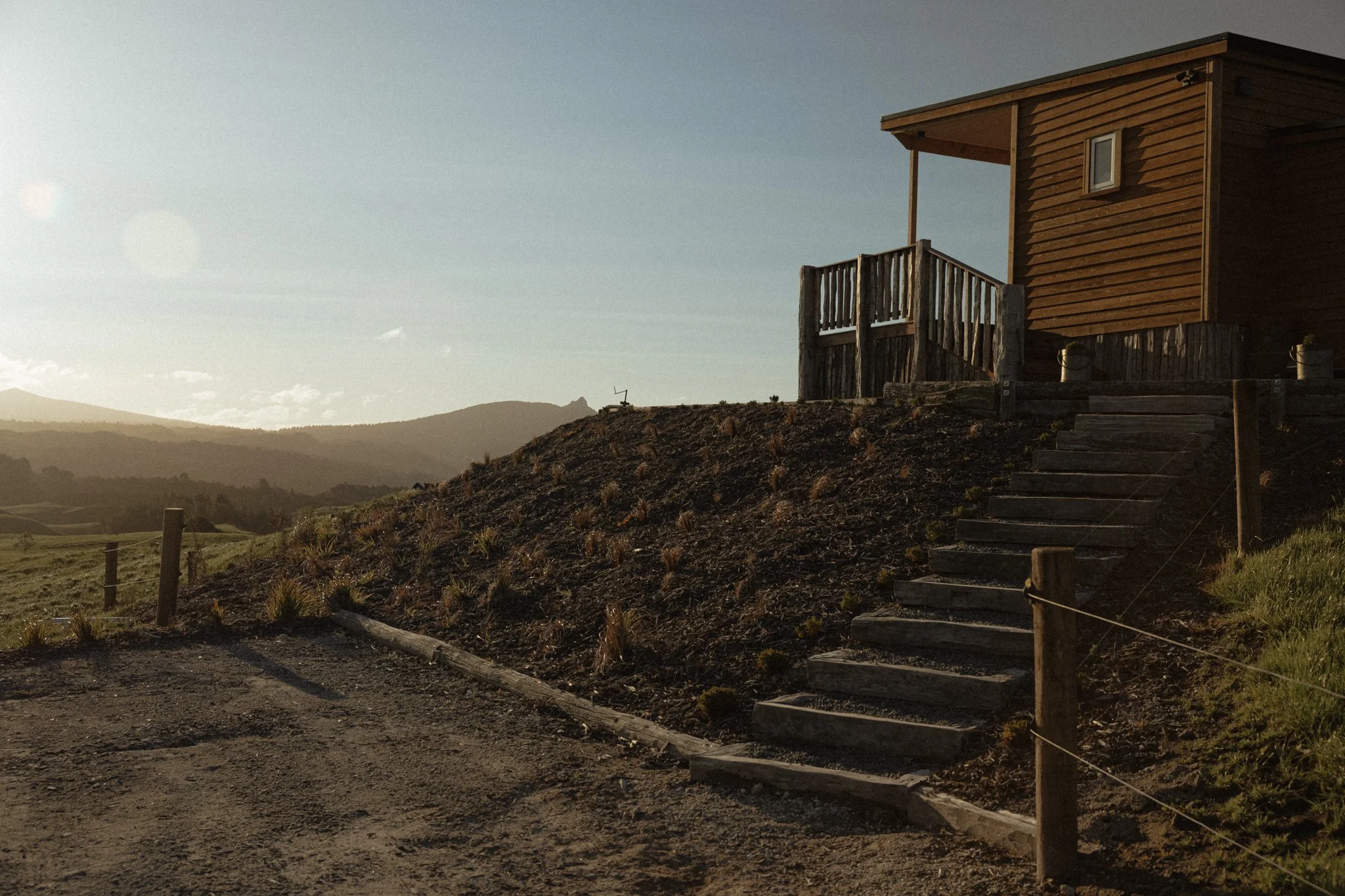A wooden cabin on a hill with stairs leading up to The Farmers Rest surrounded by open landscape and mountains in the distance during sunset.