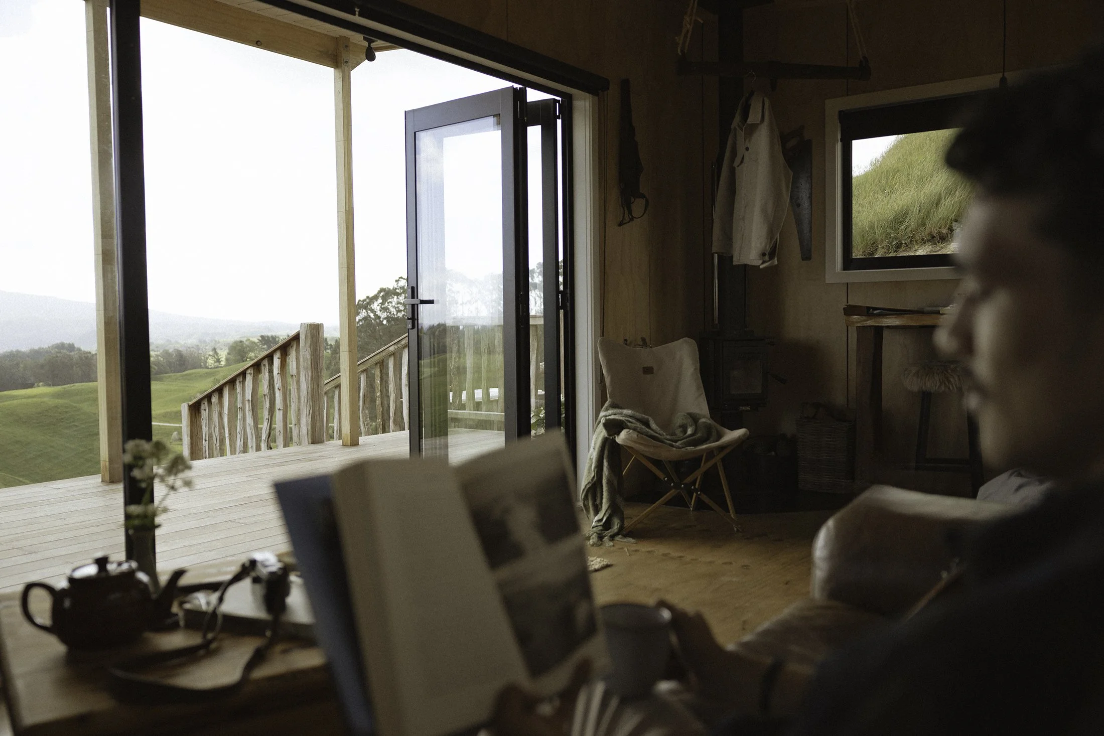 A guest sitting in a cozy interior reading a book with a cup of coffee, with an open door leading to a wooden deck and a scenic view of grassy hills and trees outside at The Farmers Rest farm stay