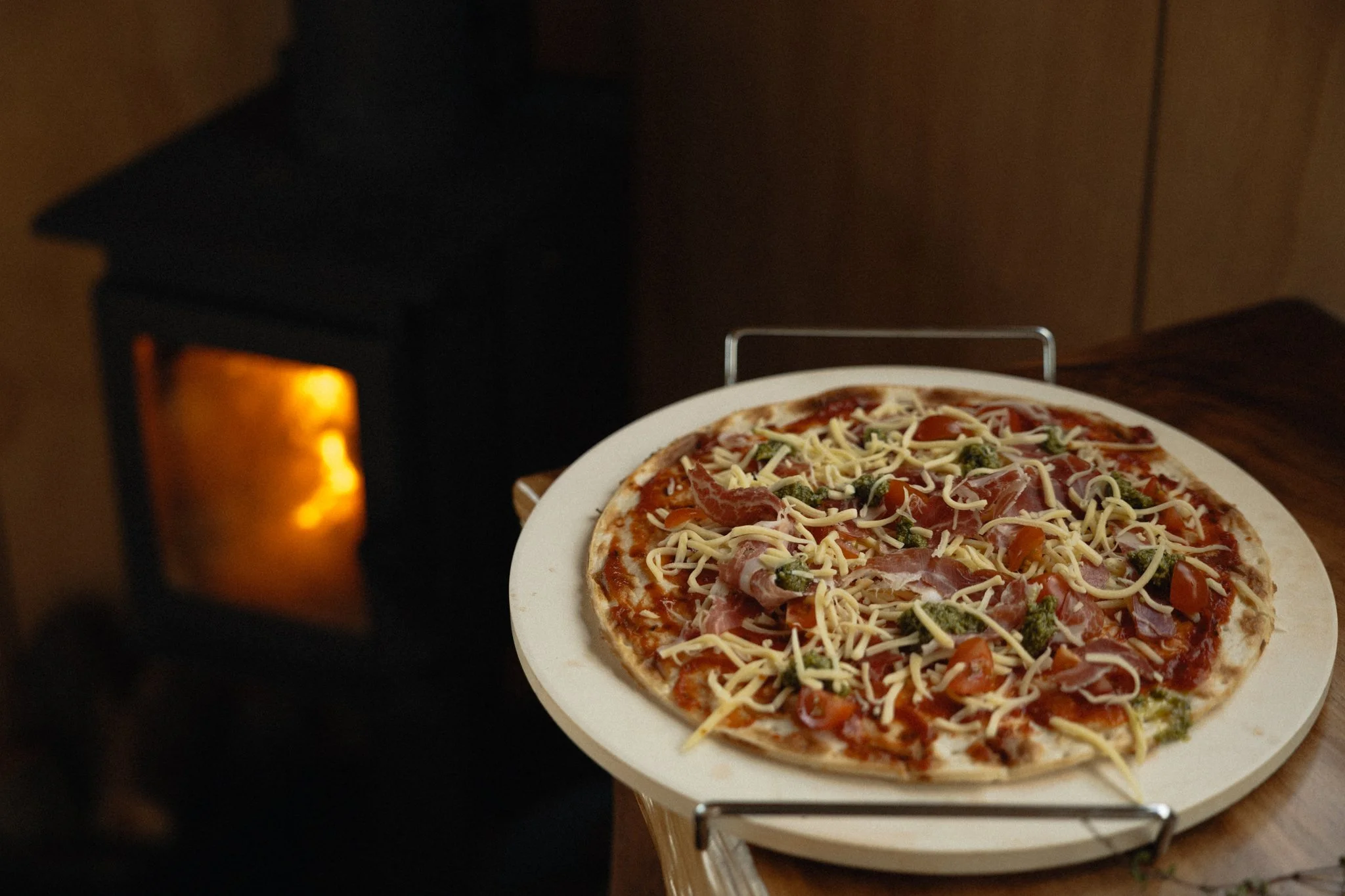 Fresh pizza with cheese, tomatoes, ham, and vegetables on a serving tray next to a lit fireplace at The Farmers Rest.
