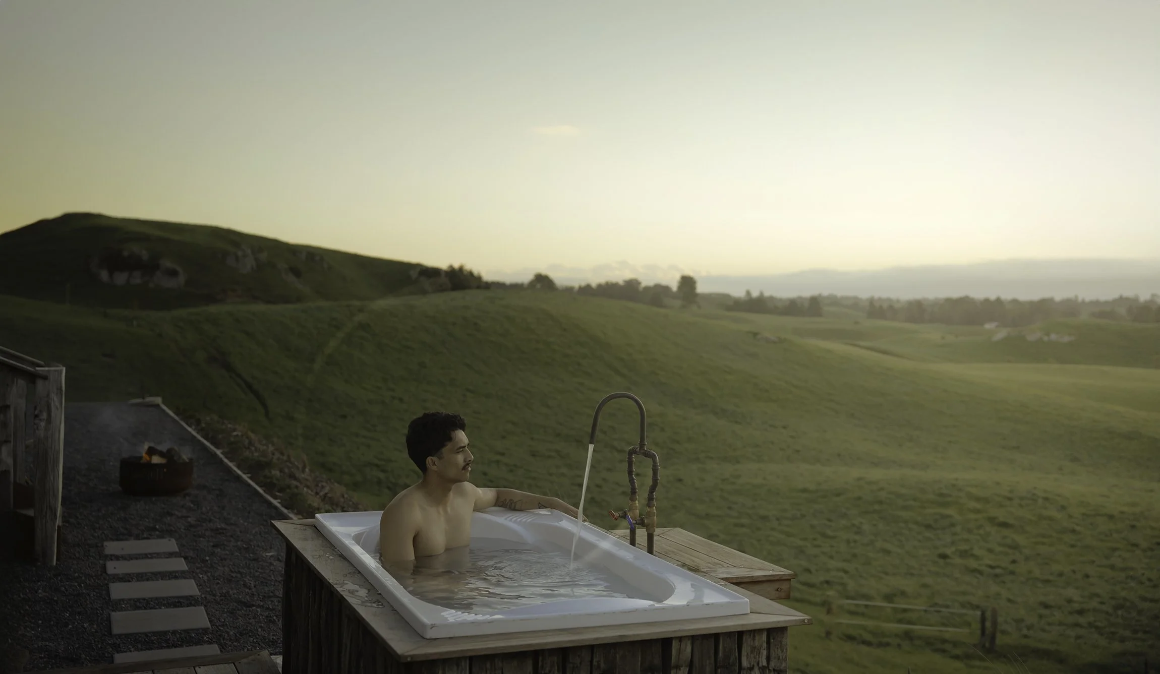 A guest at The Farmers Rest is relaxing in an outdoor bathtub on a wooden deck overlooking rolling green hills at sunset.