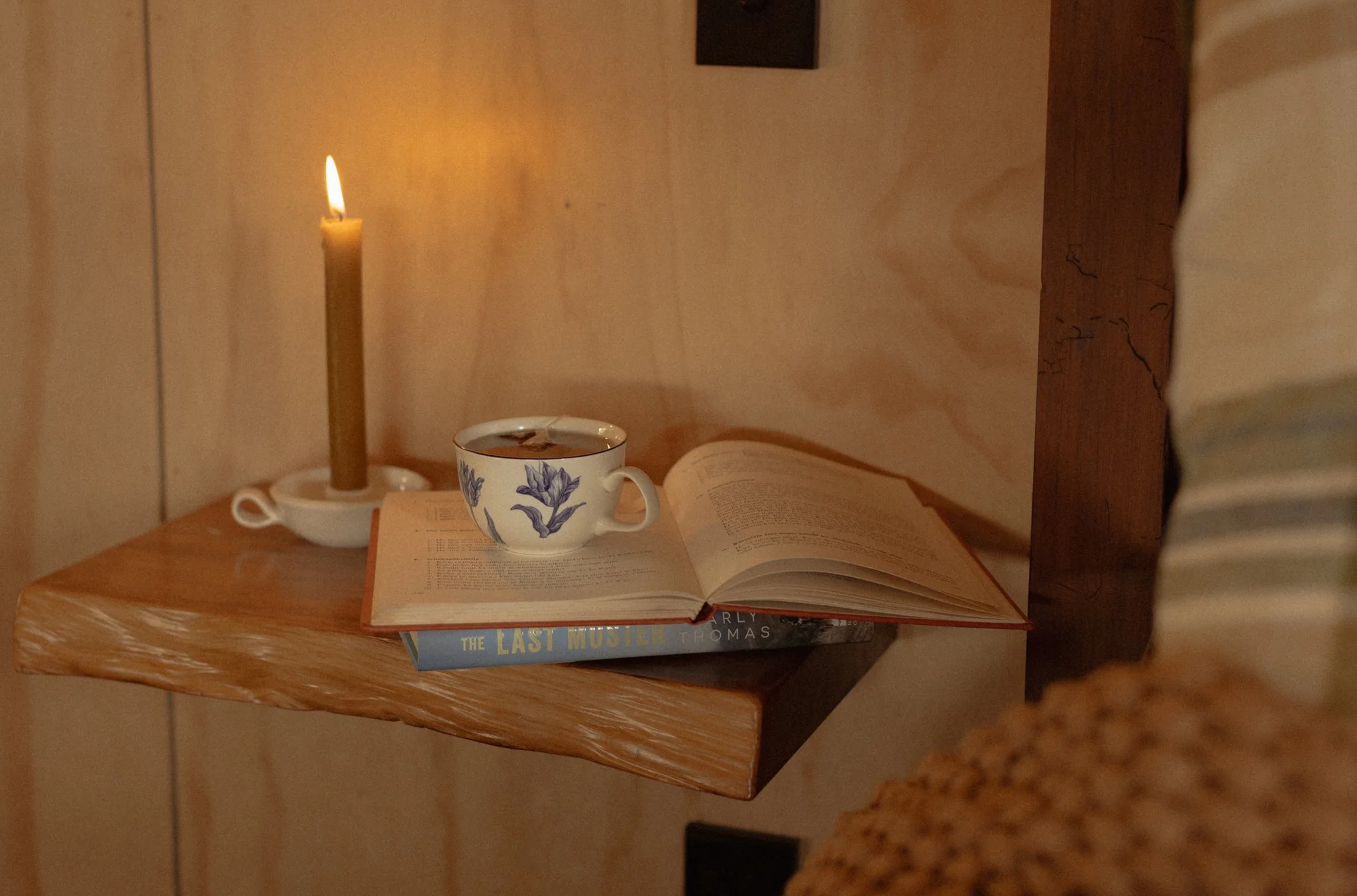 A wooden side table with an open book, a mug of tea, and a lit candle in a small holder in the lounge of The Farmers Rest private cabin stay.