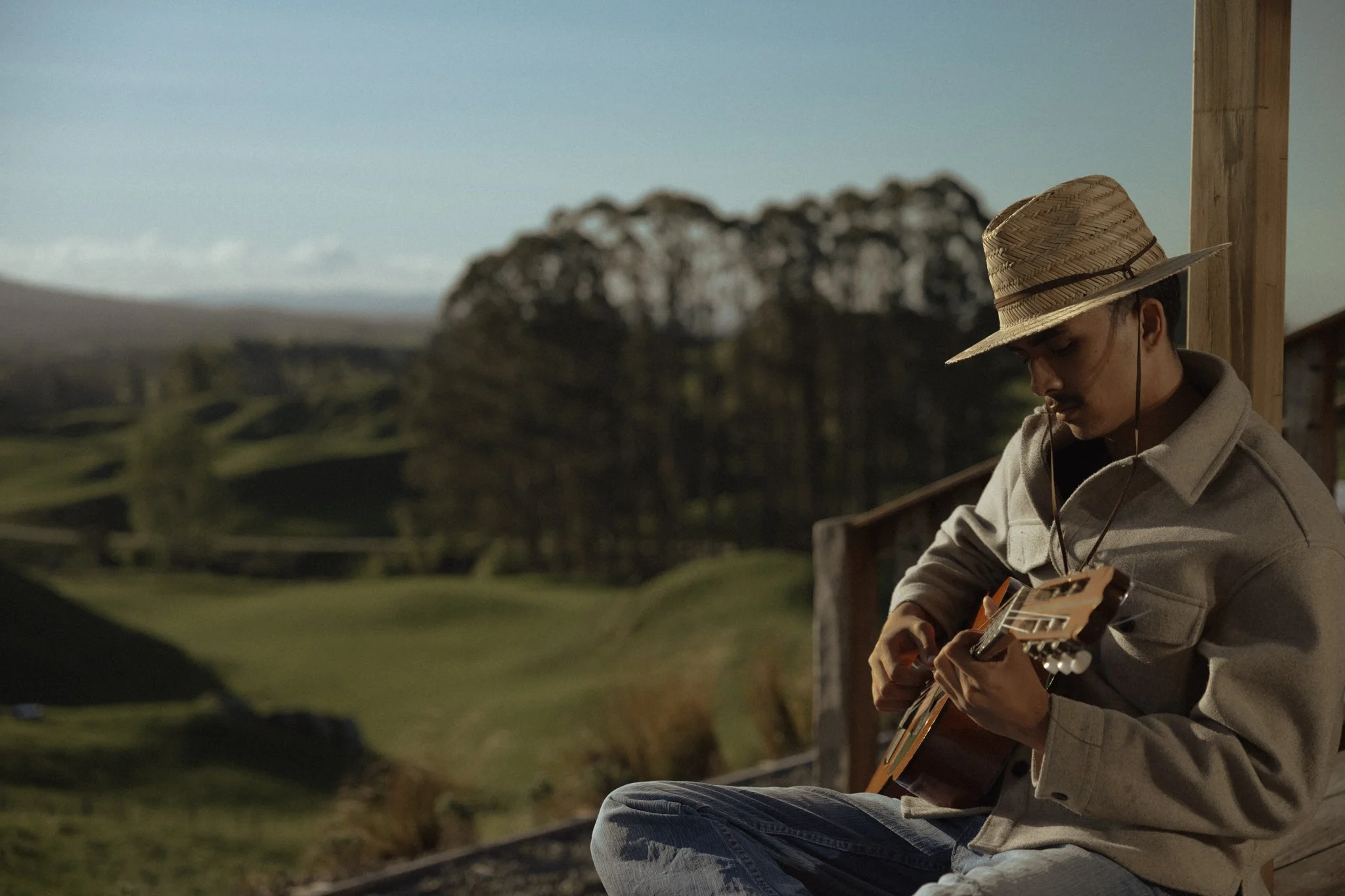 A guest sitting on a porch at The Farmers Rest wearing a straw hat and beige jacket, playing a guitar, with a scenic landscape of rolling green hills and trees in the background.