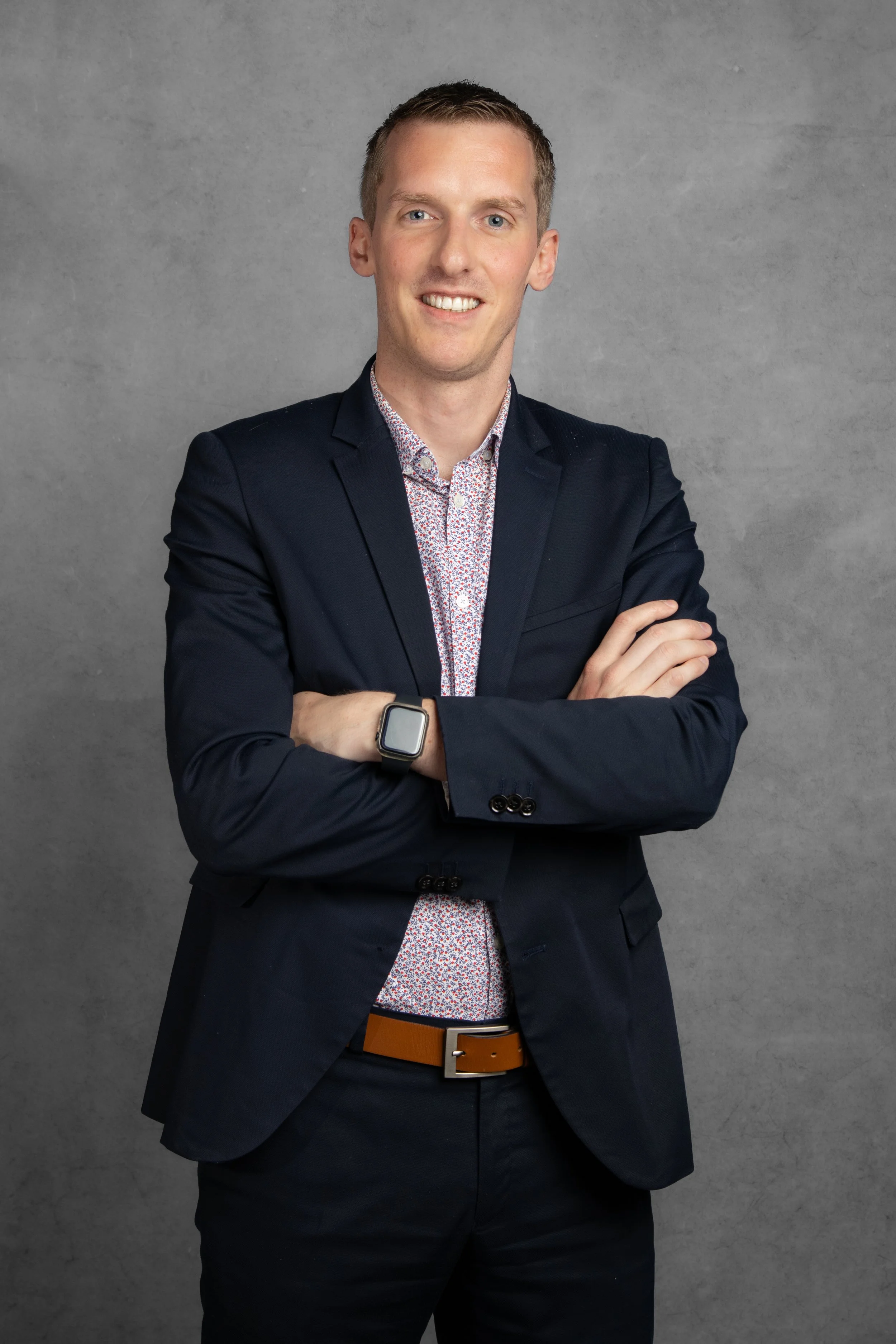A young man with short hair, wearing a dark suit and a patterned shirt, smiling with arms crossed in front of a gray textured background.