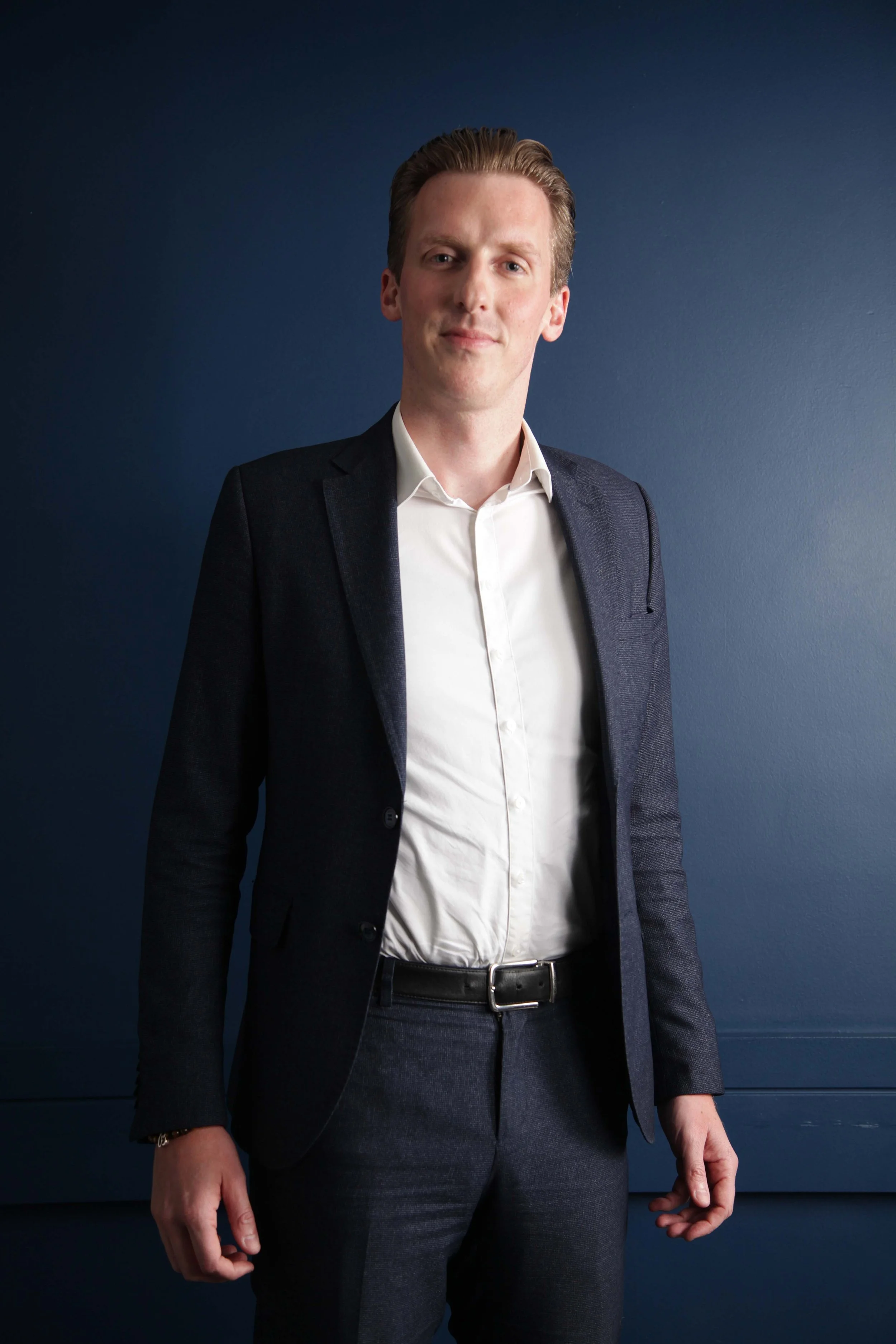 A young man with light skin, wearing a white dress shirt and a dark blazer, stands against a dark blue wall. He has short, slicked-back hair and a subtle smile.