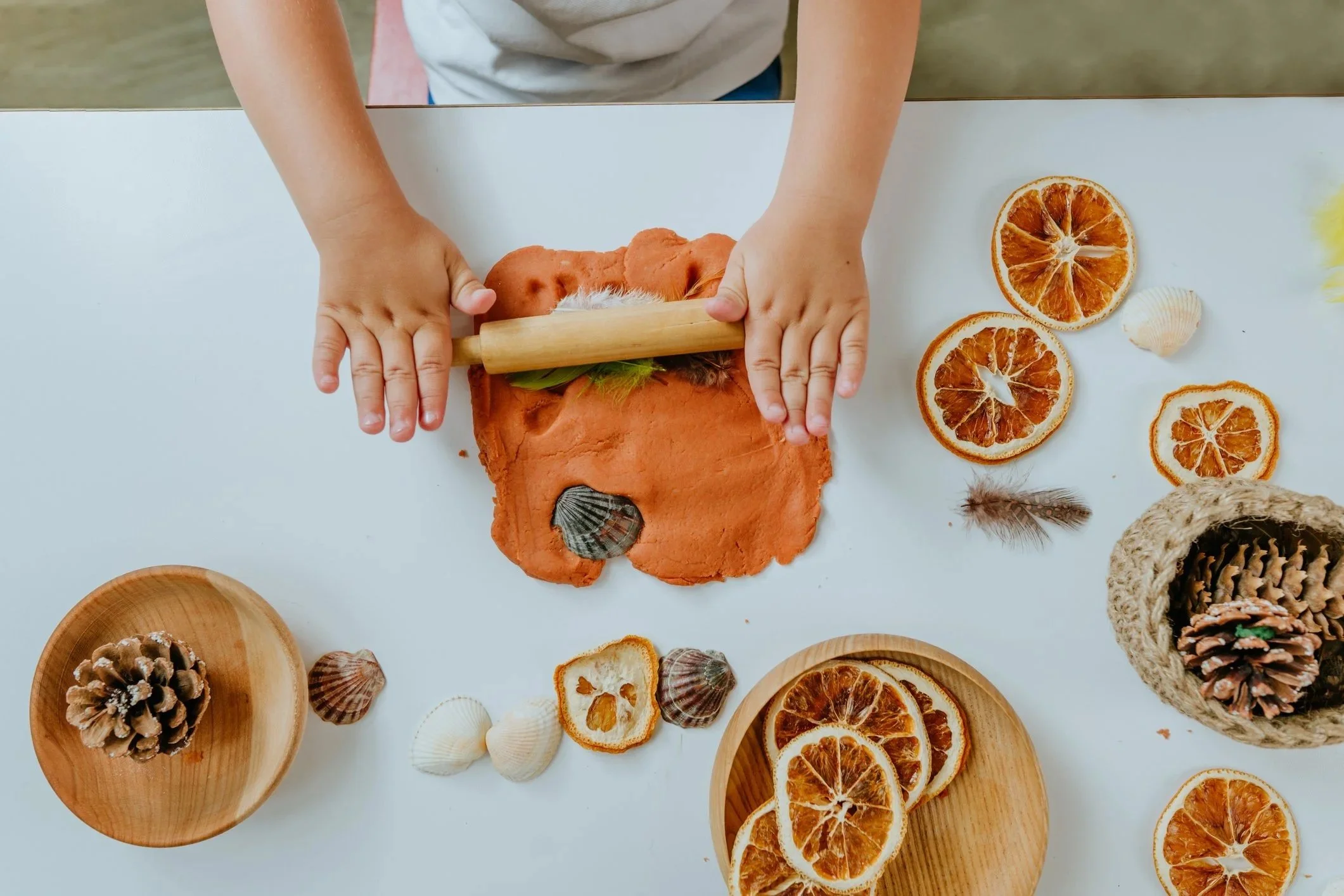 Child rolling out orange-colored clay with a wooden rolling pin, surrounded by dried orange slices, seashells, pinecones, and feathers on a white surface.