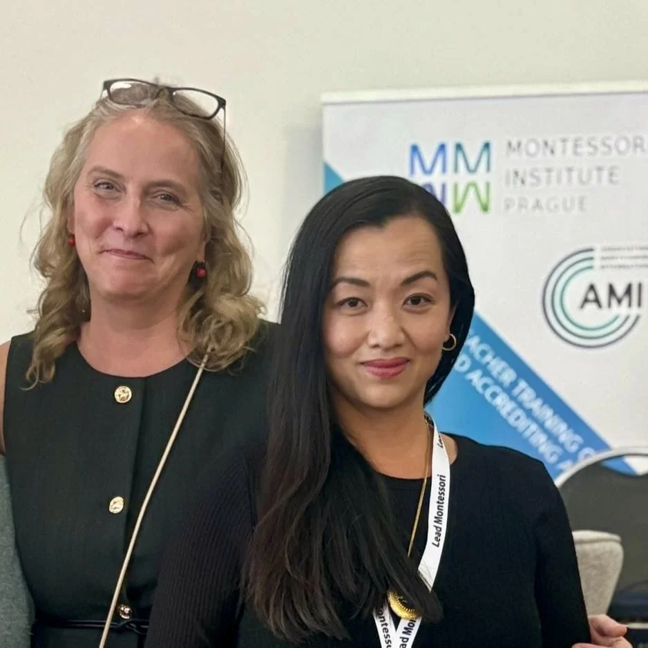 Two women standing together at an event, with a background banner displaying logos and text related to a Montessori institute and a conference.
