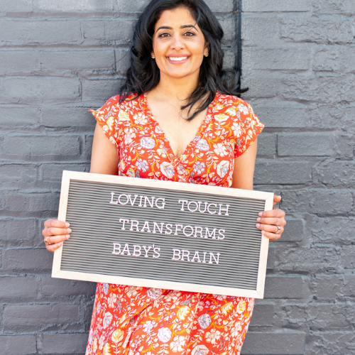 A smiling woman holding a message board that reads "Loving touch transforms baby's brain" in front of a gray brick wall.