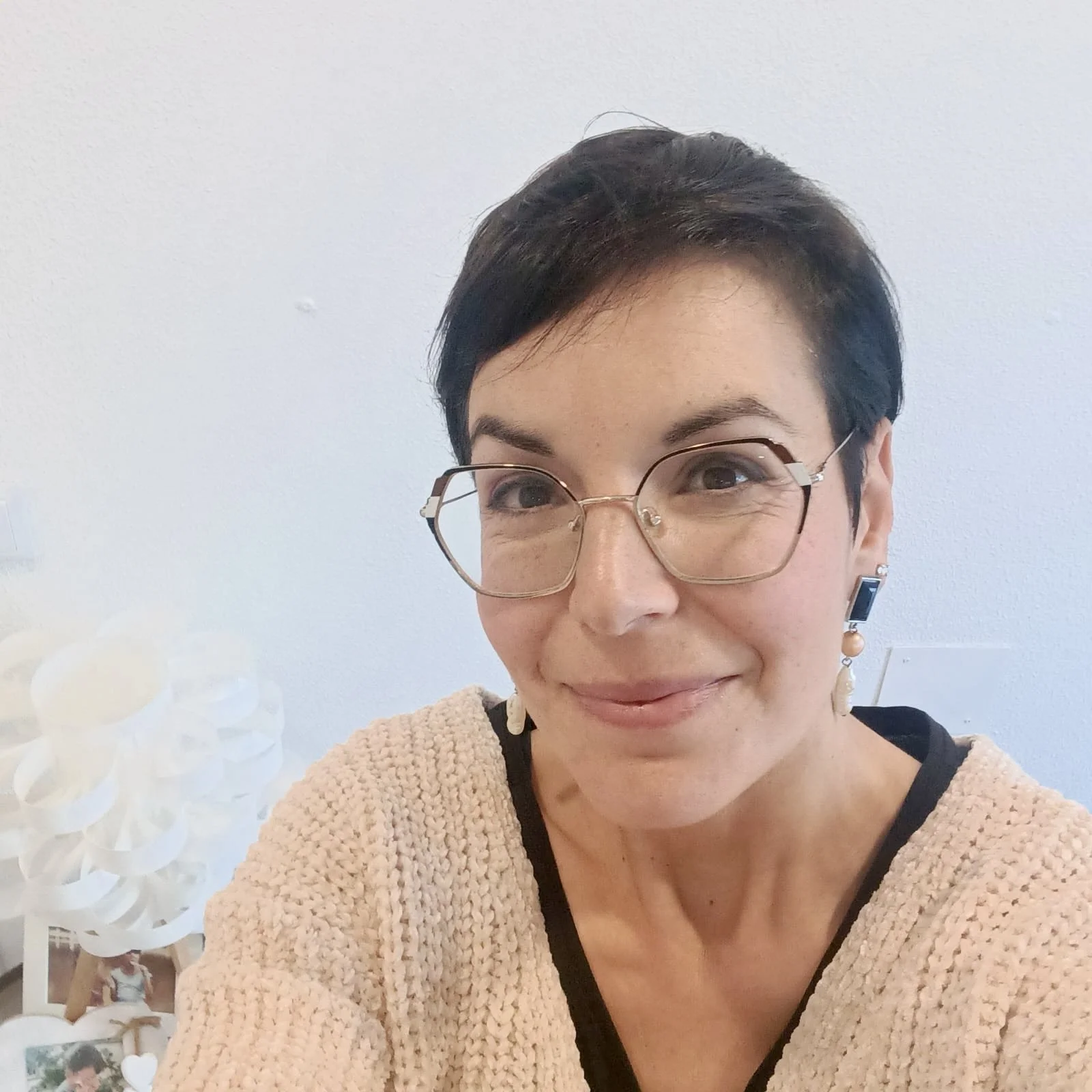 A woman with short dark hair and glasses smiling indoors, wearing earrings and a beige top.
