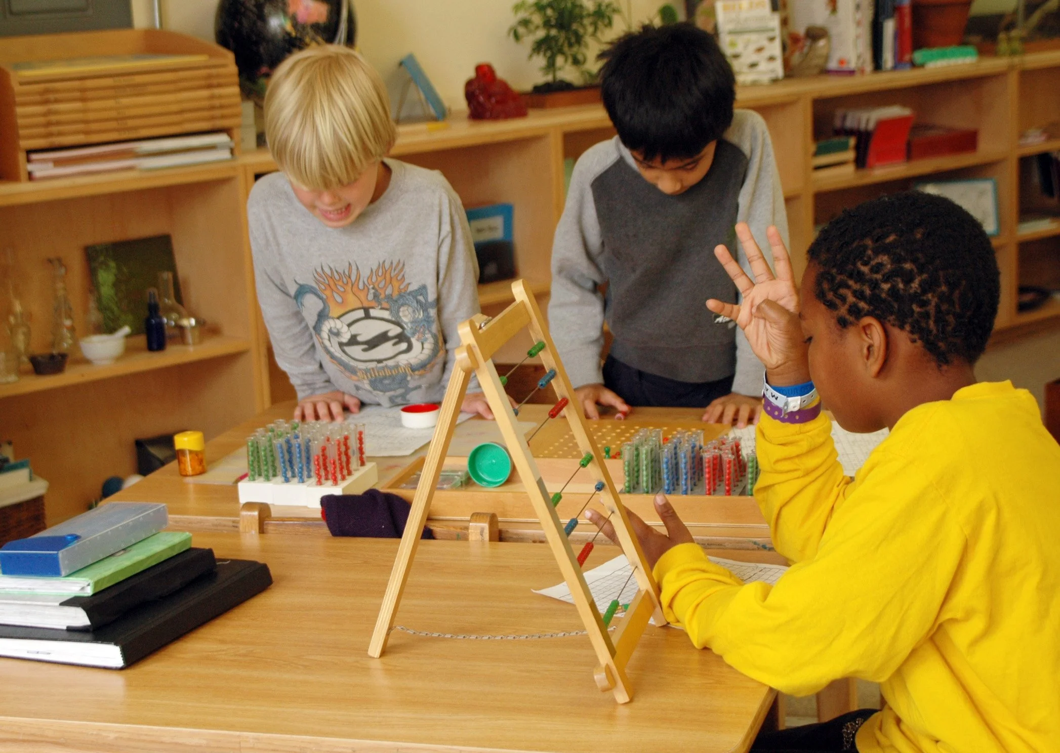 Three children in a classroom playing with a homemade abacus and colorful counting beads. One child with dark hair and wearing a yellow jacket is showing a hand gesture, while the other two children, one with blond hair and the other with black hair, are looking at the abacus.