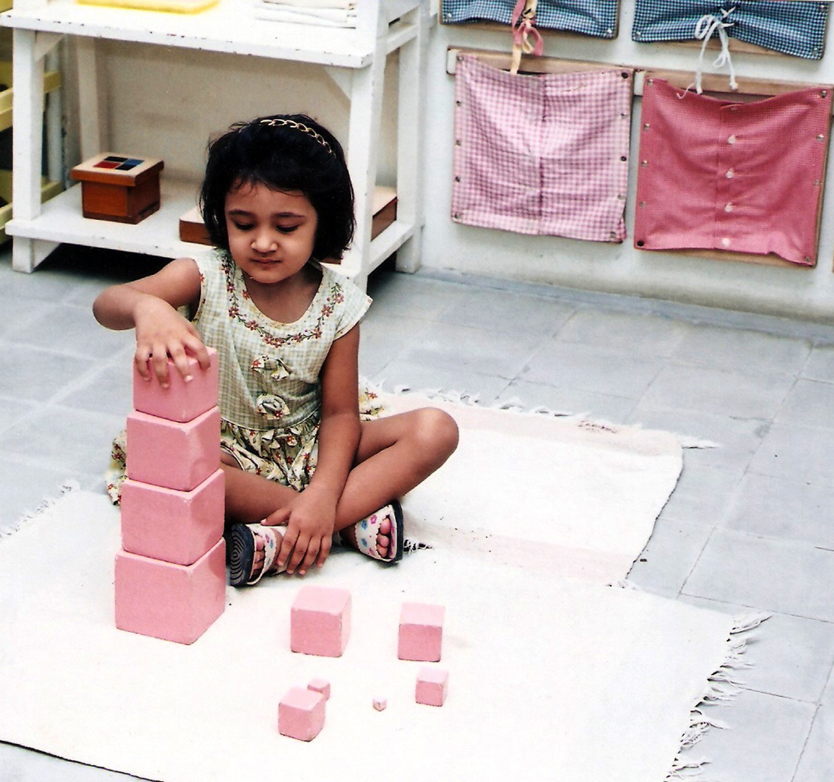 A young girl sitting cross-legged on a white rug, stacking pink foam blocks into a tower of five, with additional blocks of various sizes arranged in front of her, in a room with a tiled floor and fabric-covered wall organizers.