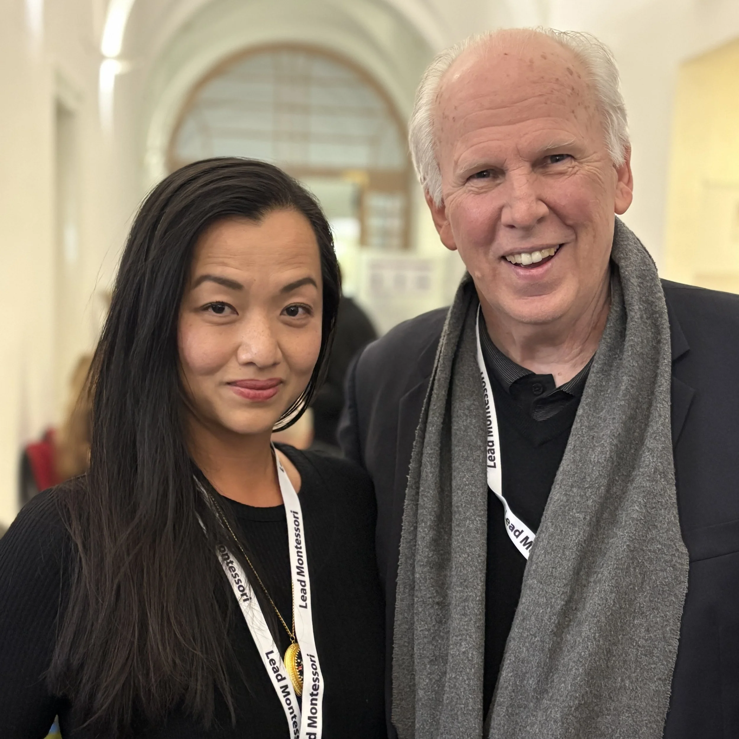 A woman and a man smiling and posing for a photo at an indoor event, wearing conference lanyards that say 'Lead Montessori.'