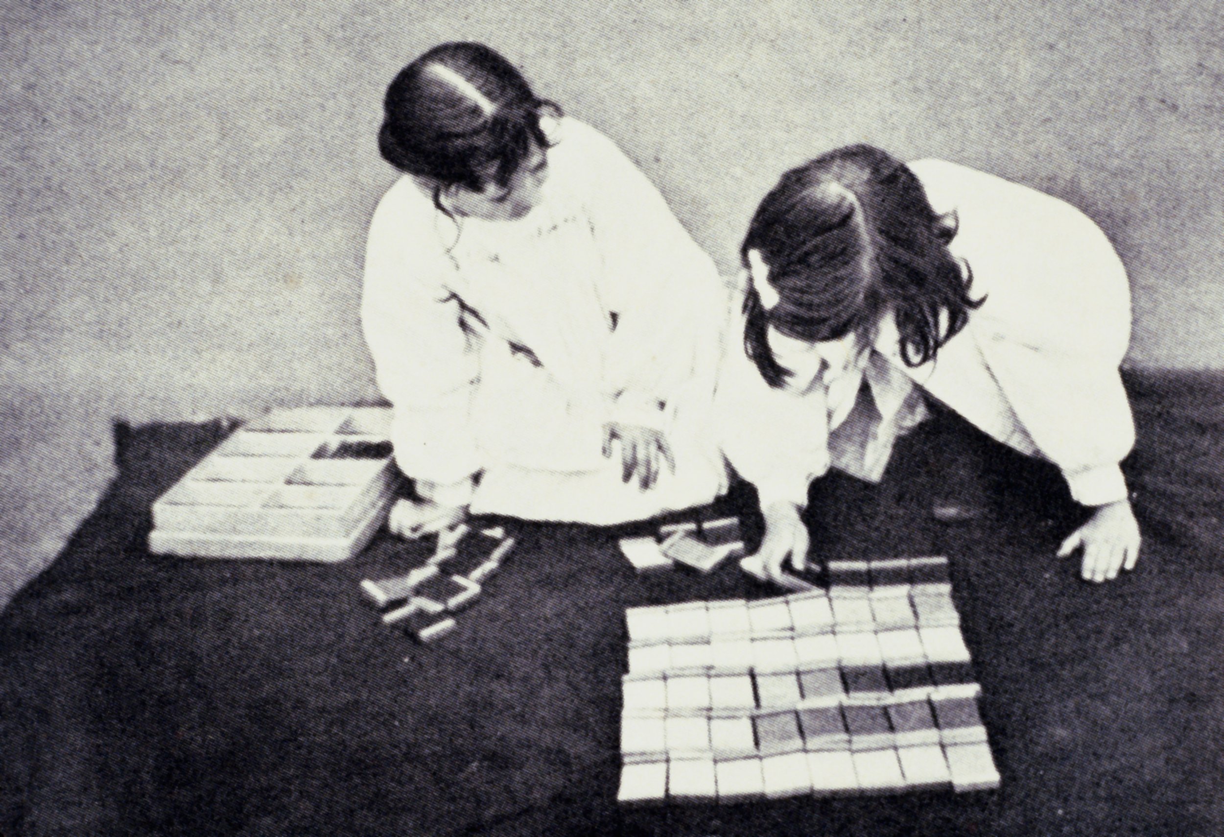Two girls are sitting on the floor playing a tile game with multiple stacks of tiles in front of them.