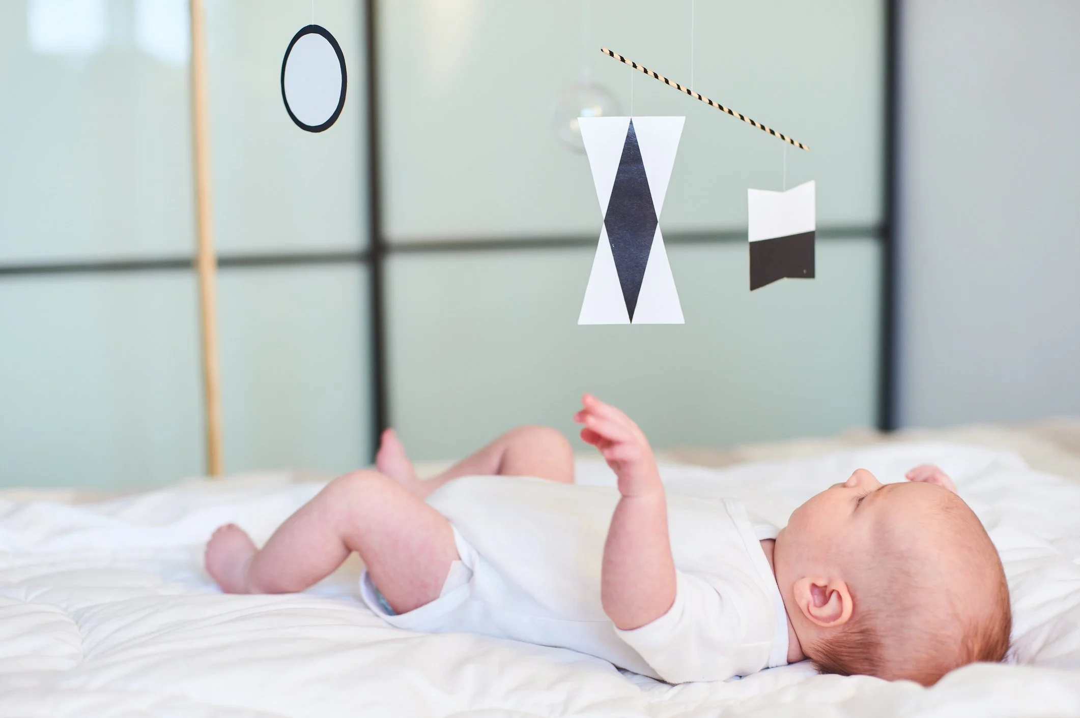 A baby lying on a white bed looking at hanging geometric black-and-white decorations, with a light green wall in the background.